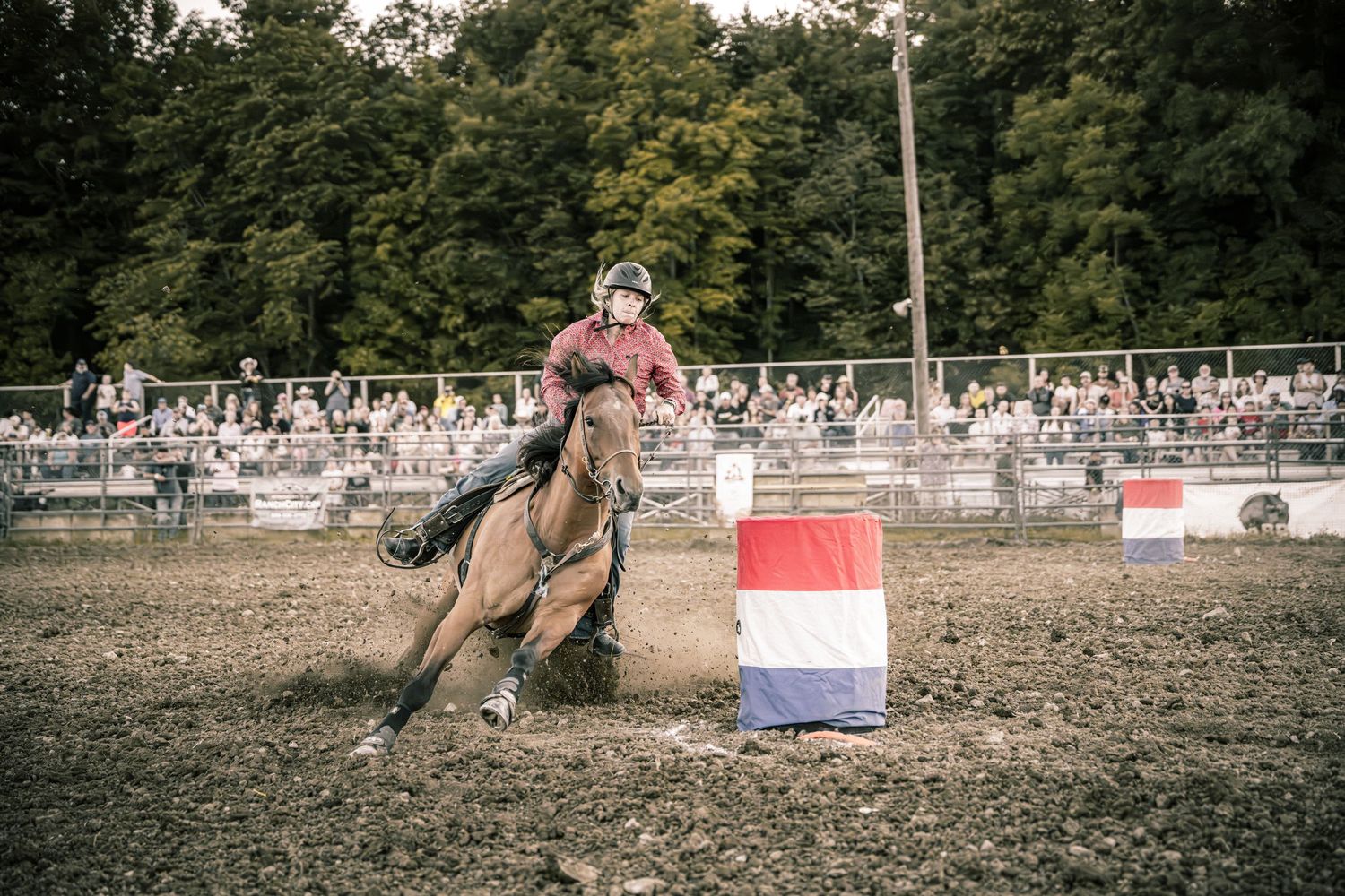 Barrel racing cowboy on a brown horse rounding a red, white, and blue barrel at a rodeo.