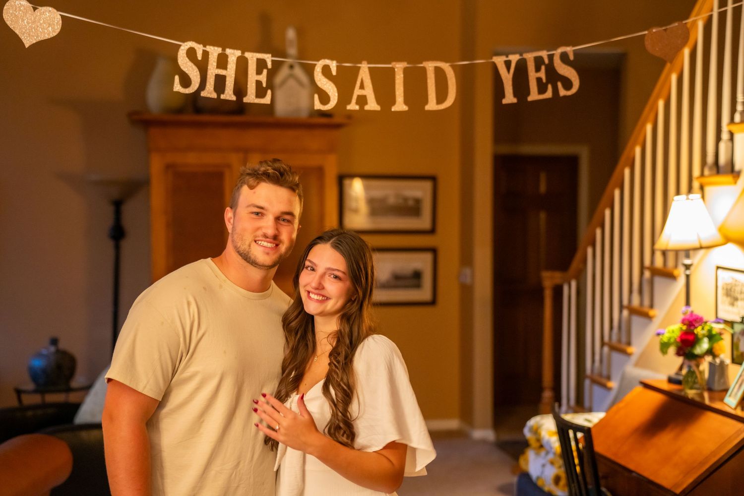 Newly engaged couple smiling indoors under a 'She Said Yes' banner after a proposal.