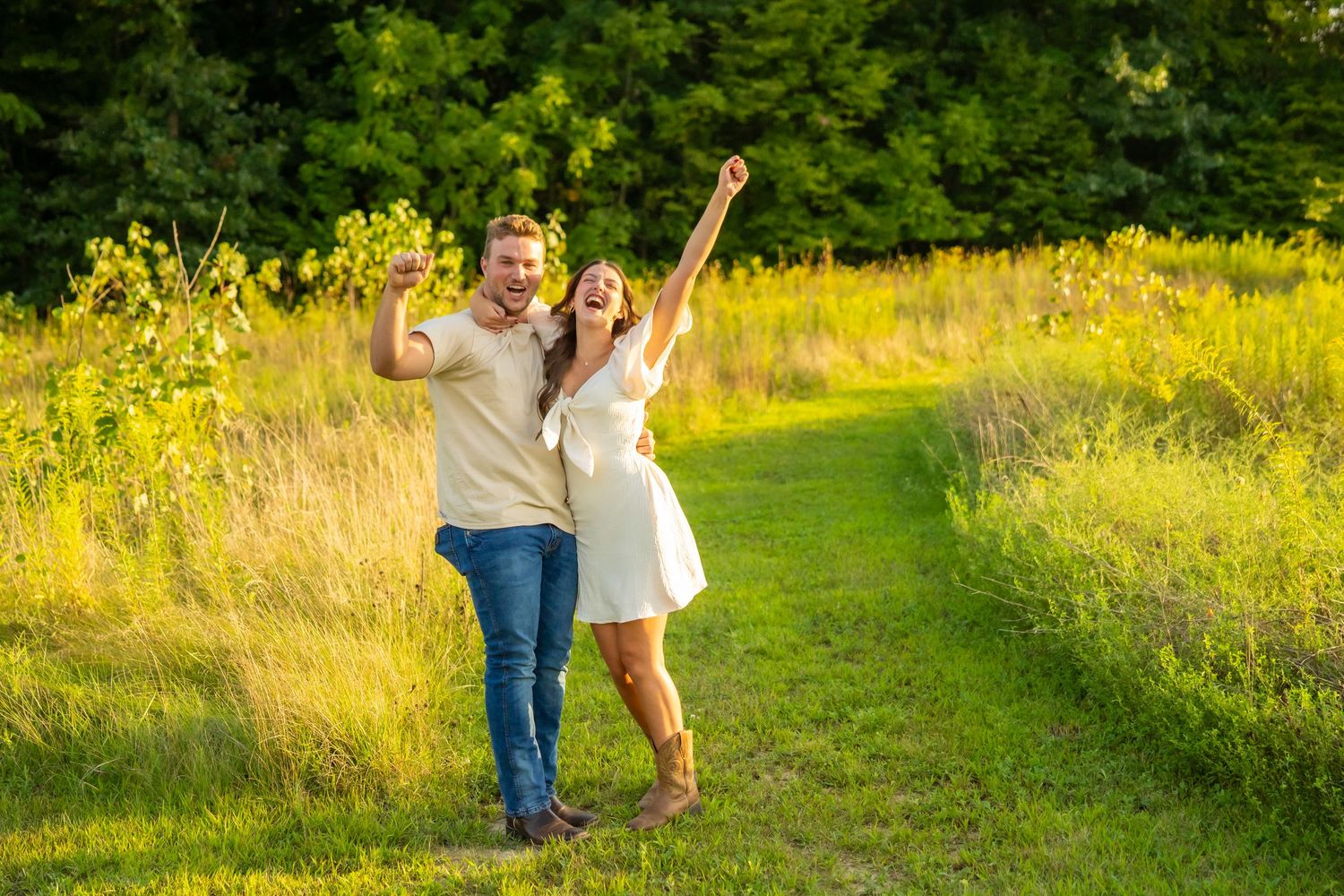 Joyful newly engaged couple celebrating with raised arms on a sunny green meadow path.