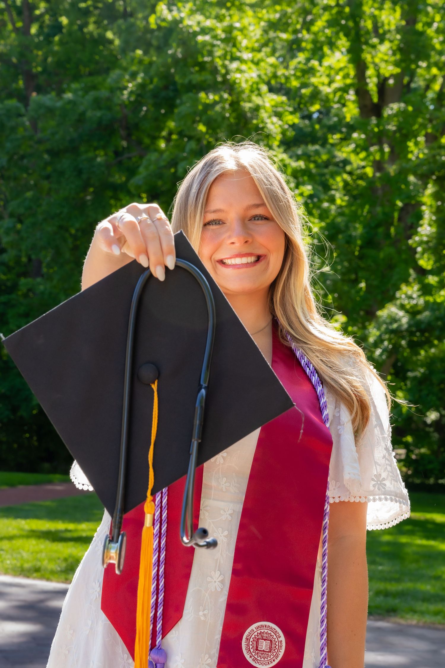 Happy blonde graduate holding mortarboard cap with stethoscope and red graduation stole outdoors.