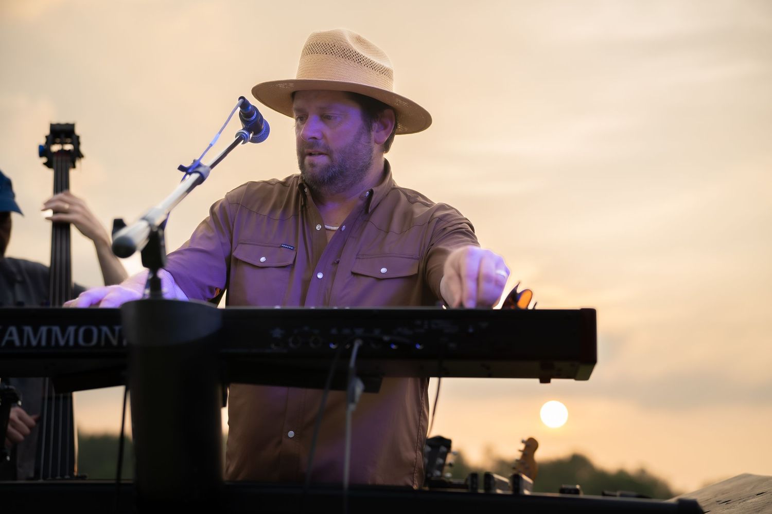Male musician playing Hammond organ keyboard at outdoor sunset concert wearing wide-brim hat.