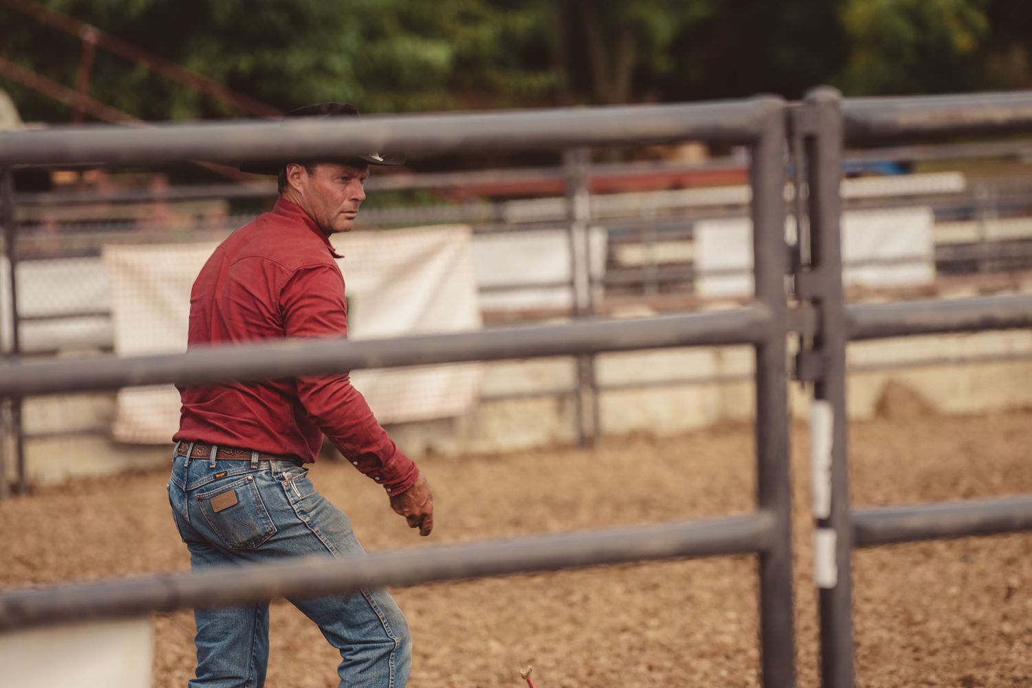 Cowboy in red shirt and jeans walking along metal livestock fence at outdoor rodeo arena.