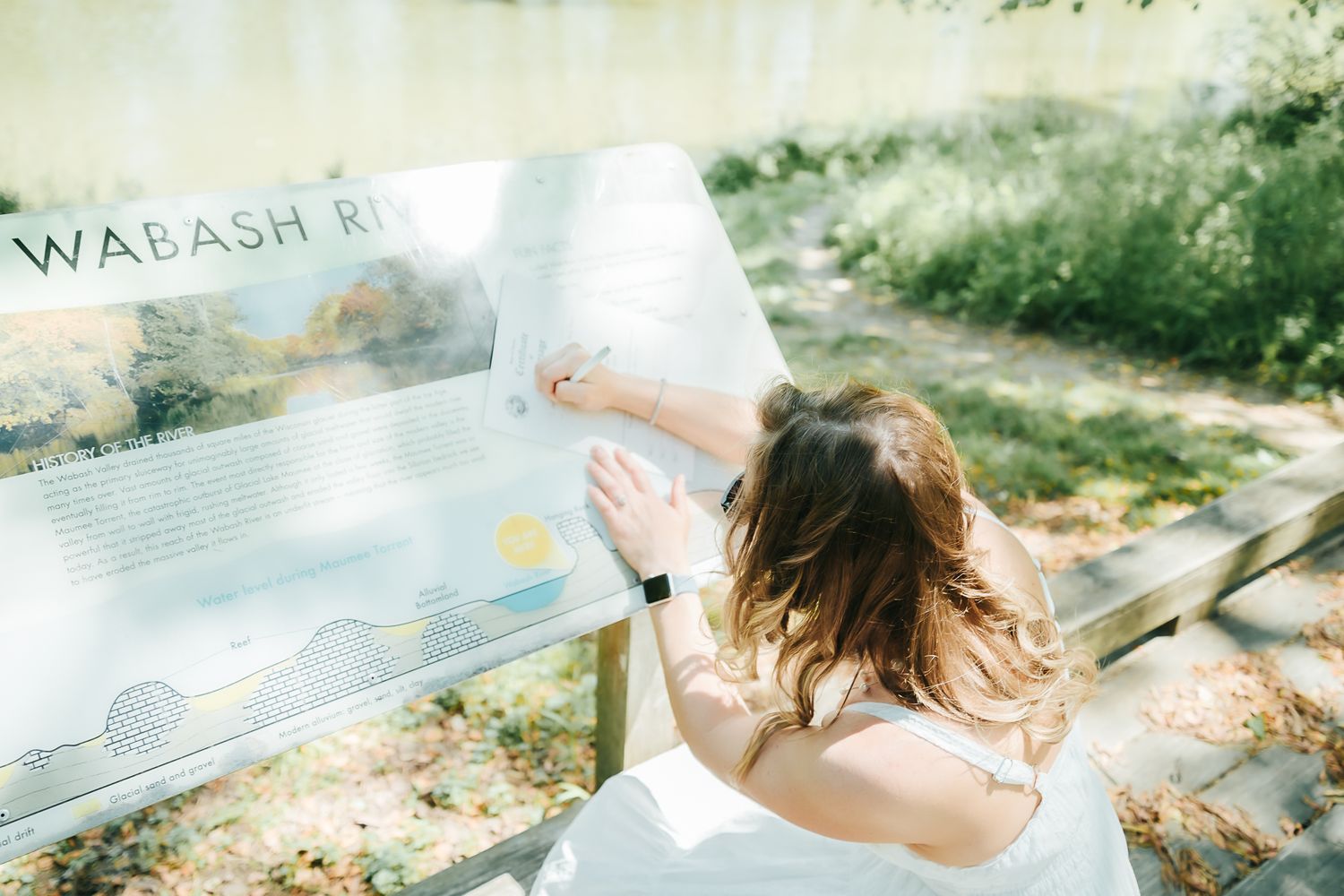 Woman in white dress reading a Wabash River informational sign outdoors near the water.