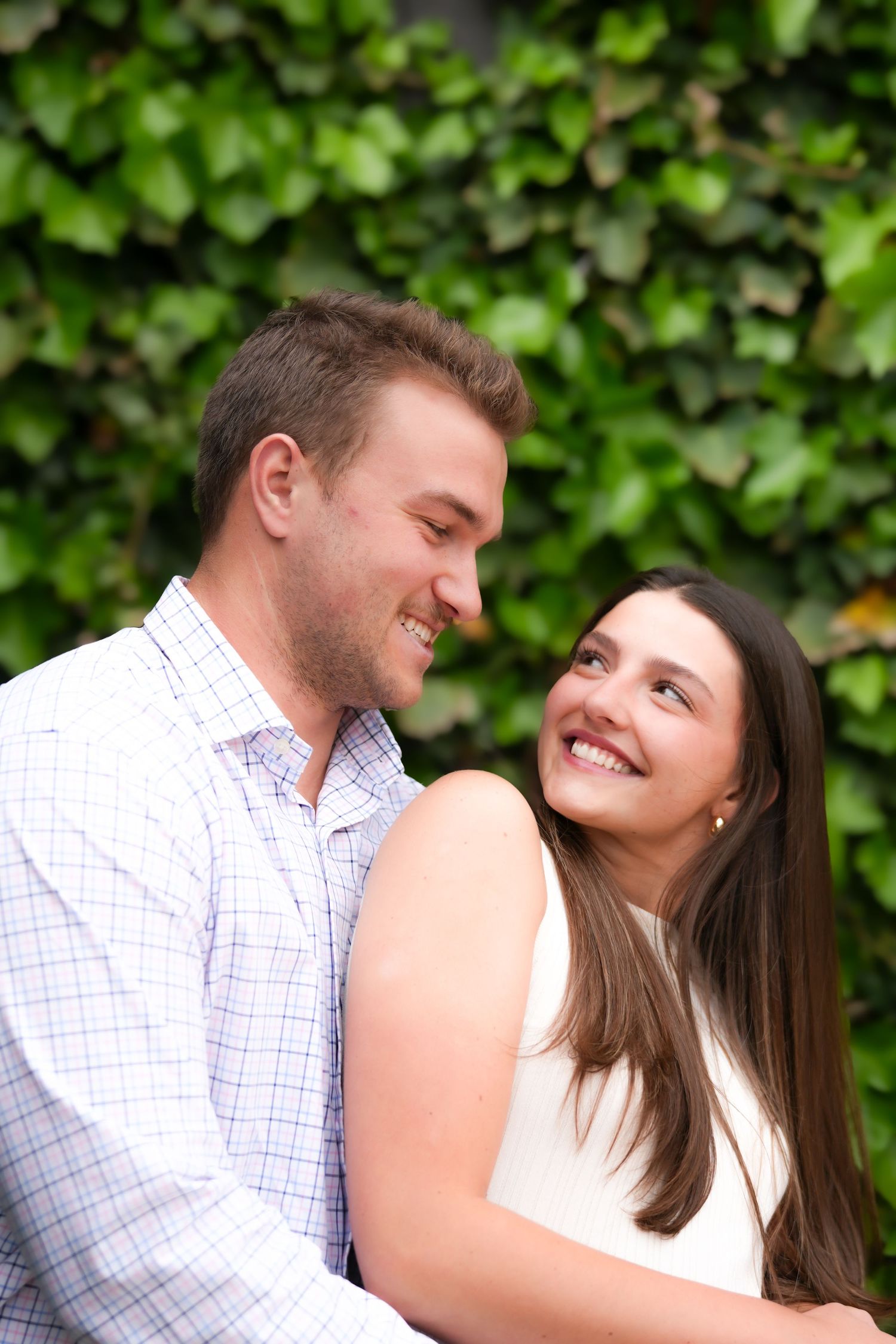 A couple shares a romantic moment smiling at each other against a lush green ivy wall.
