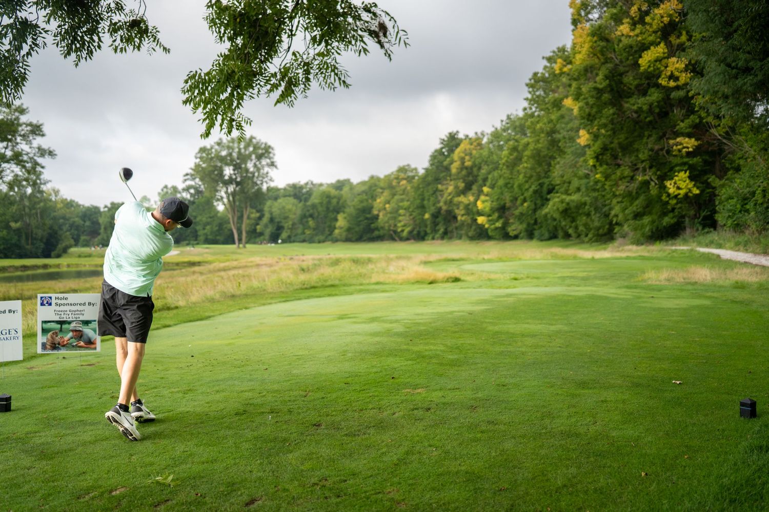 Golfer teeing off on a scenic green golf course surrounded by dense trees on a cloudy day.