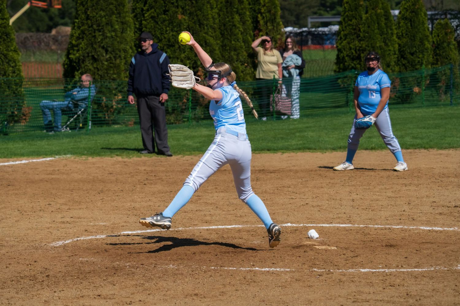 Softball pitcher in light blue uniform winds up to throw during a sunny outdoor game with teammates and spectators visible.
