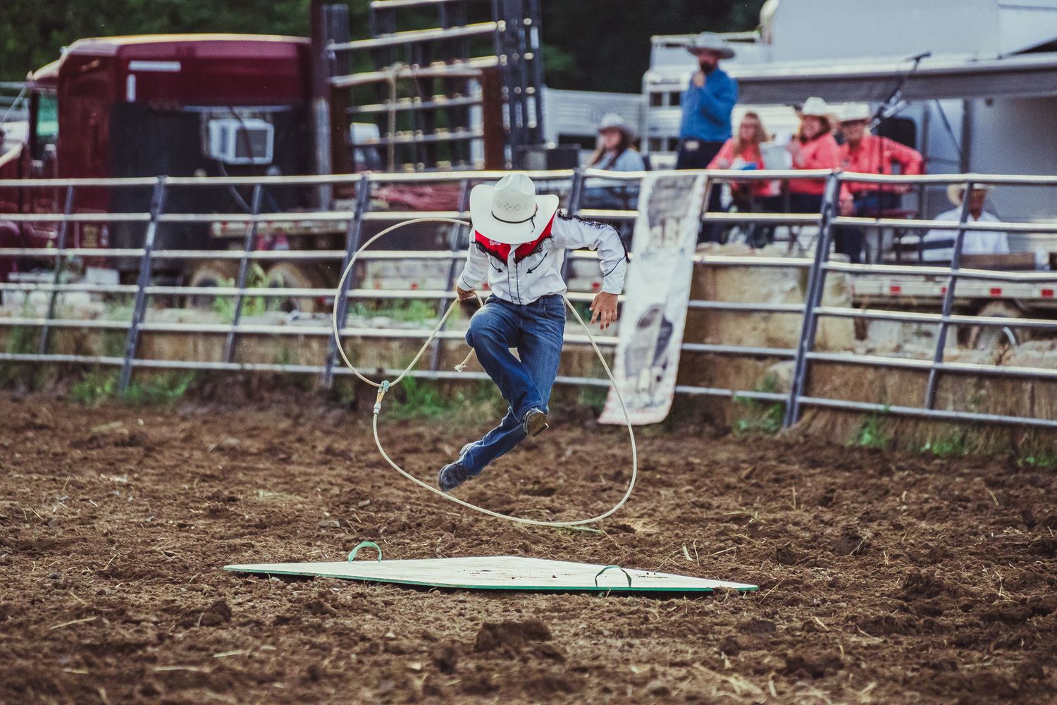 Cowboy performing rope trick jumping through lasso hoop at outdoor rodeo arena competition.