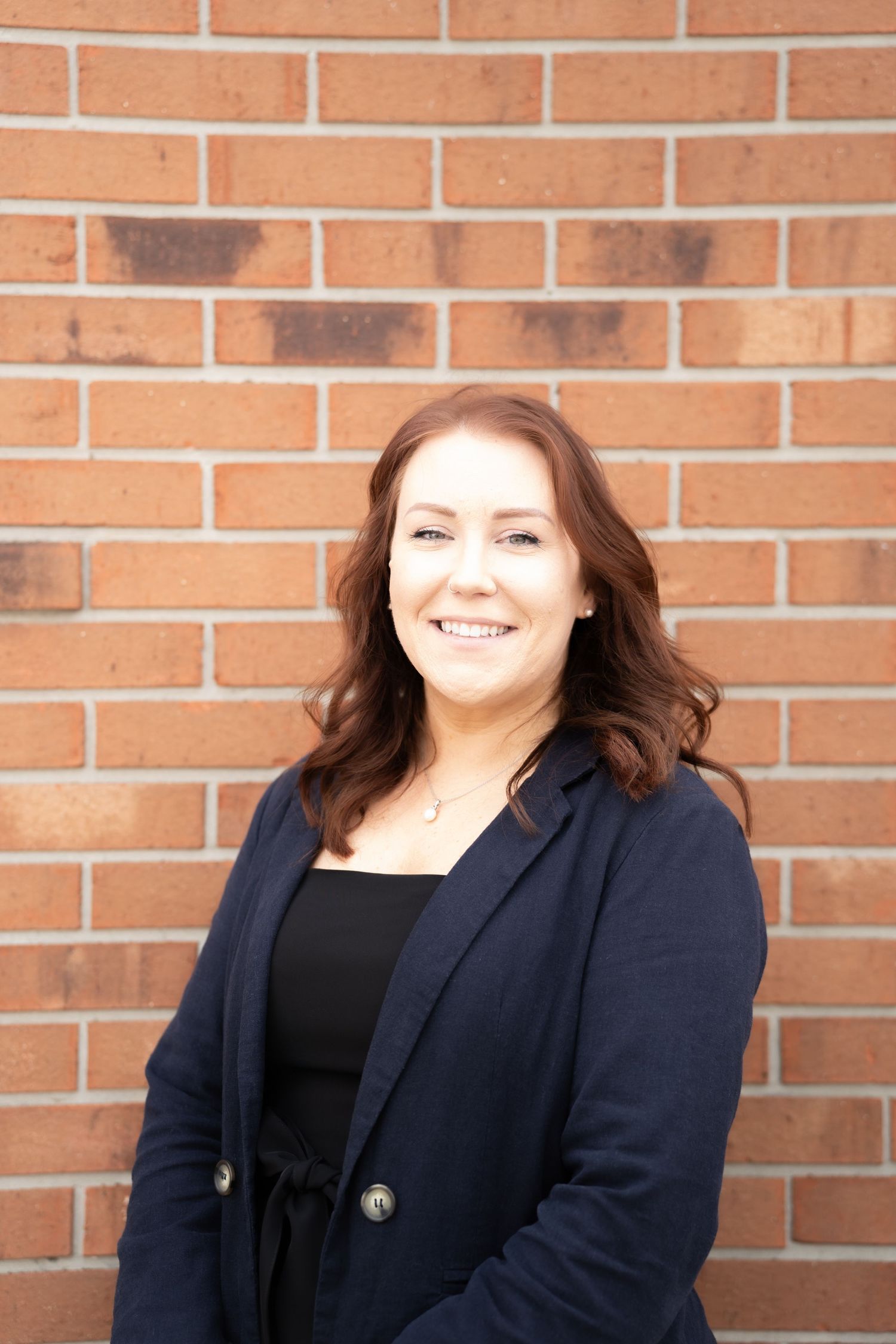 Professional woman in navy blazer smiling confidently against a red brick wall background.