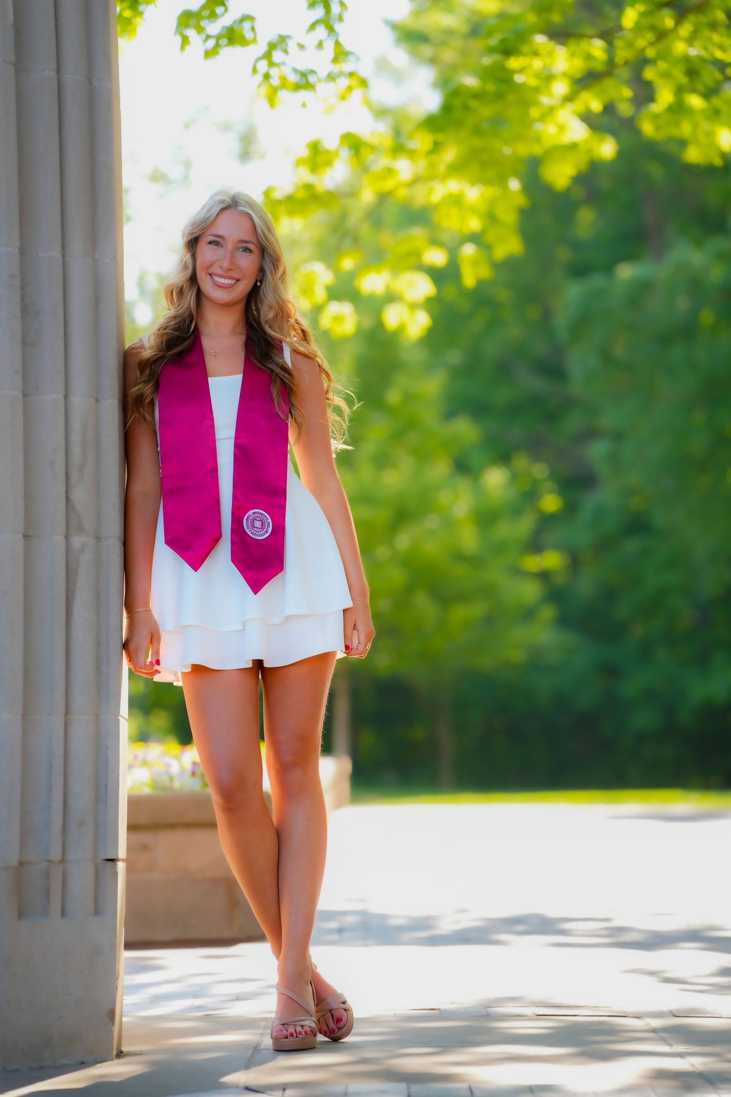 Smiling blonde graduate in white dress wearing crimson Indiana University stole poses by stone column.