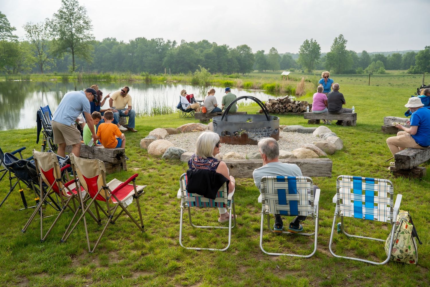 Community members relaxing in lawn chairs around a stone fire pit beside a serene pond at an outdoor gathering.