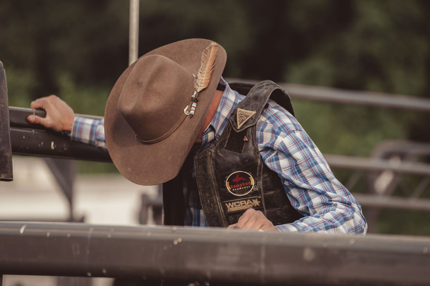 Bull rider in plaid shirt and leather vest leaning over rodeo chute before ride.
