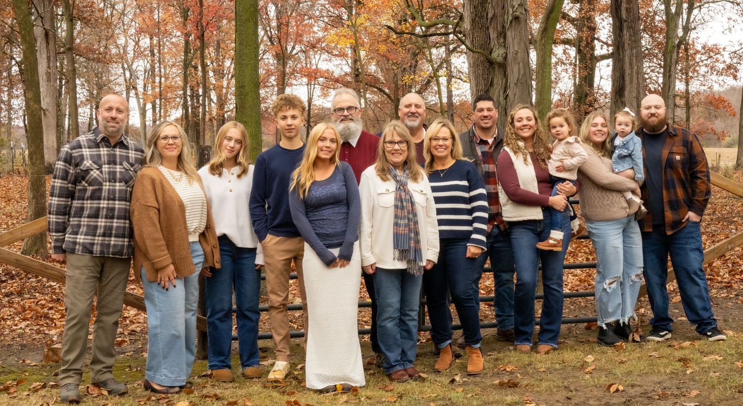 A large extended family group poses together among colorful fall foliage trees for an outdoor portrait.