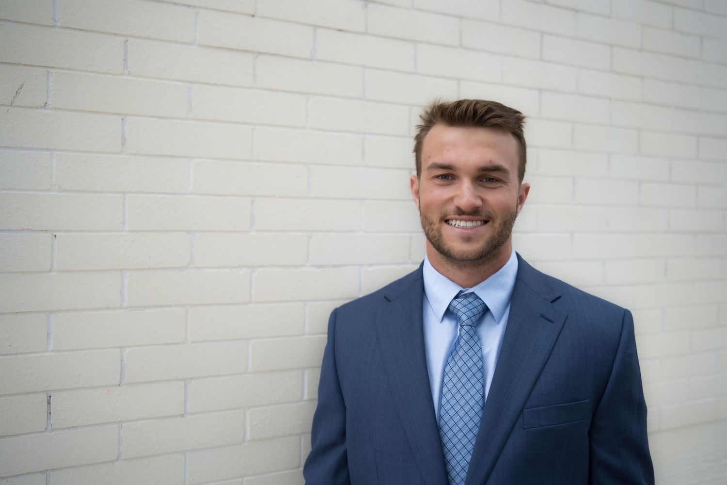 Smiling young businessman in blue suit and tie posing confidently against a white brick wall.