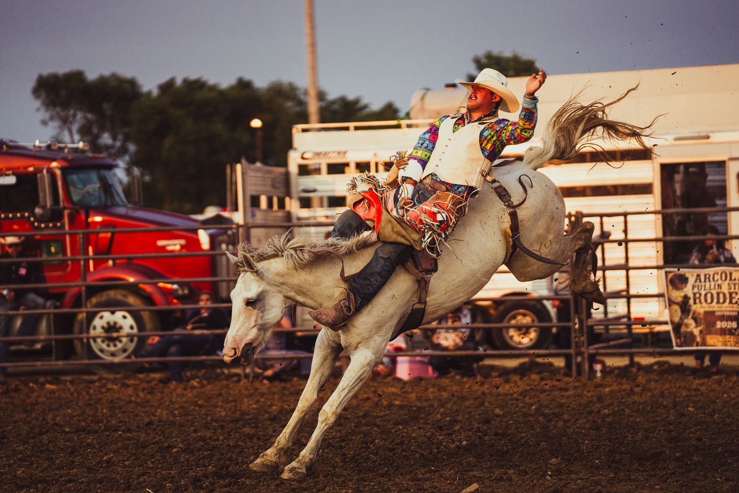 Cowboy riding a bucking white horse at a rodeo competition during golden hour.