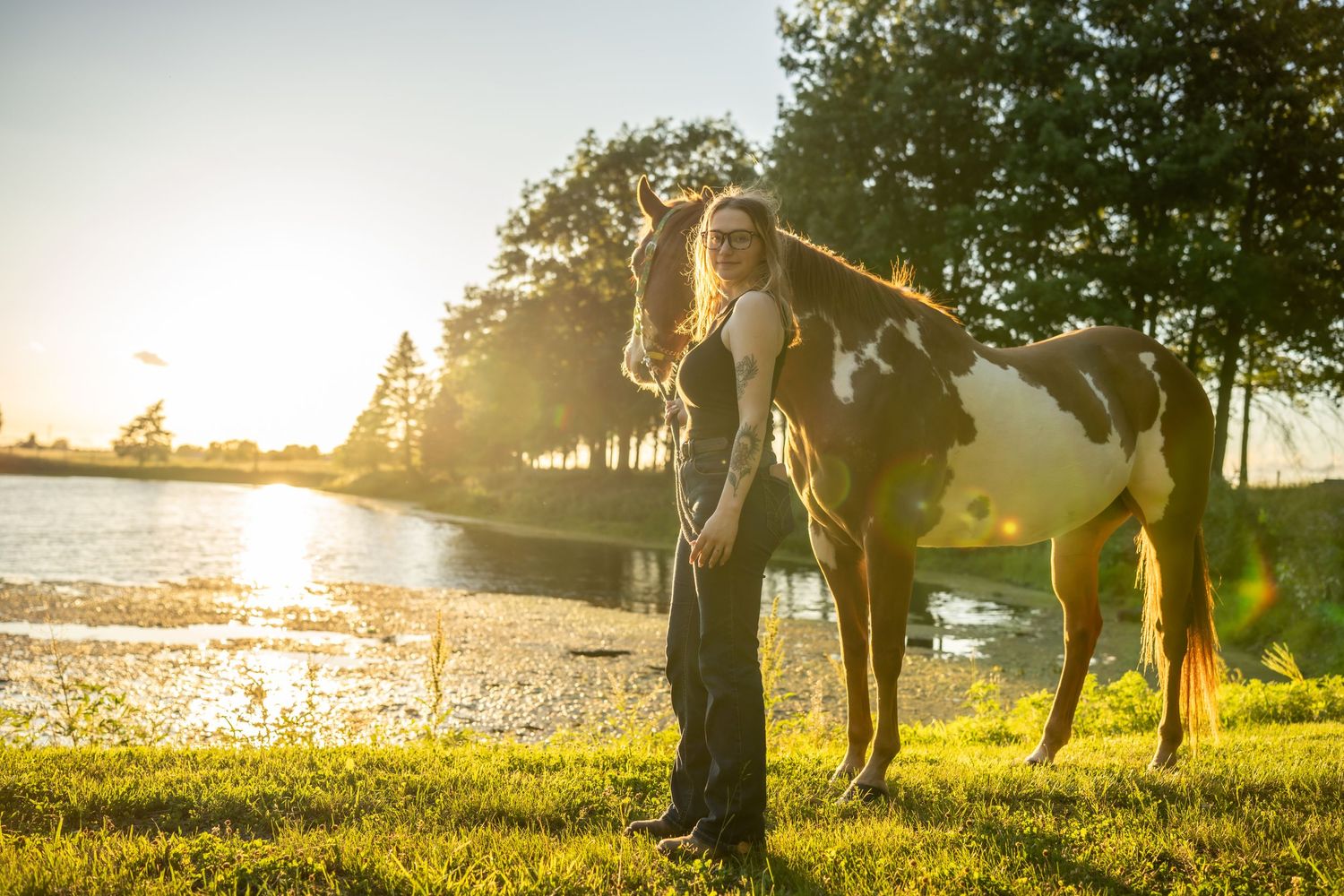 Woman standing beside a paint horse near a sunlit lake at golden hour on lush green grass.
