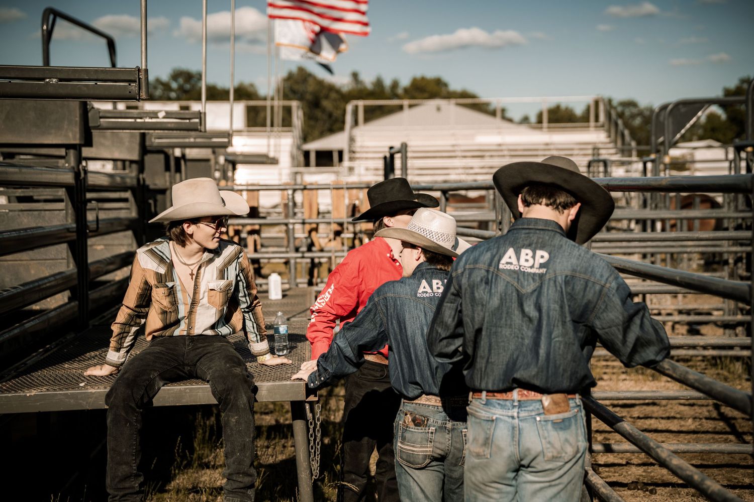 Young cowboys in hats and ABP vests gather by metal rodeo chutes with an American flag waving in the background.