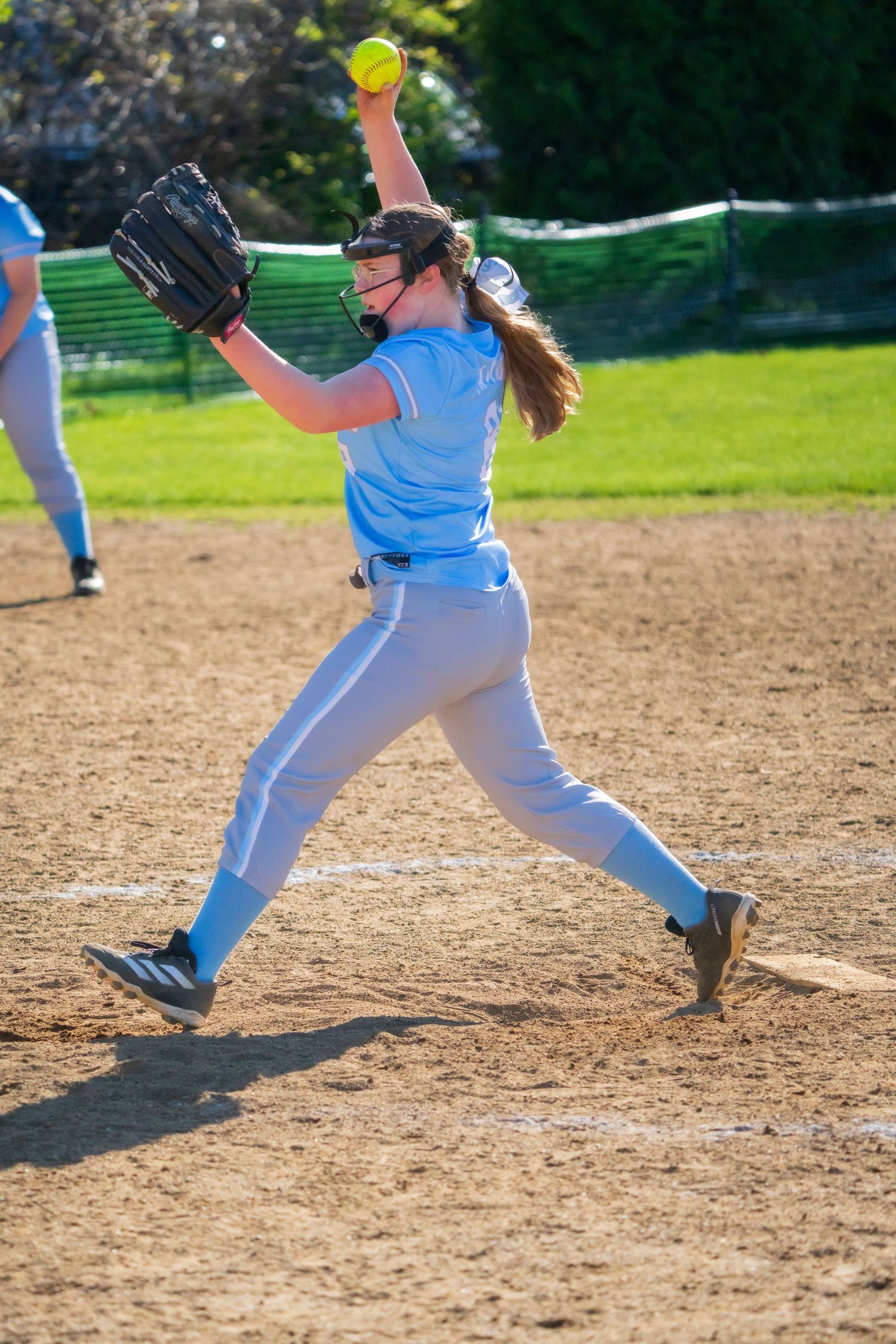 Female softball pitcher in blue uniform mid-windup, releasing yellow ball during a sunny outdoor game.
