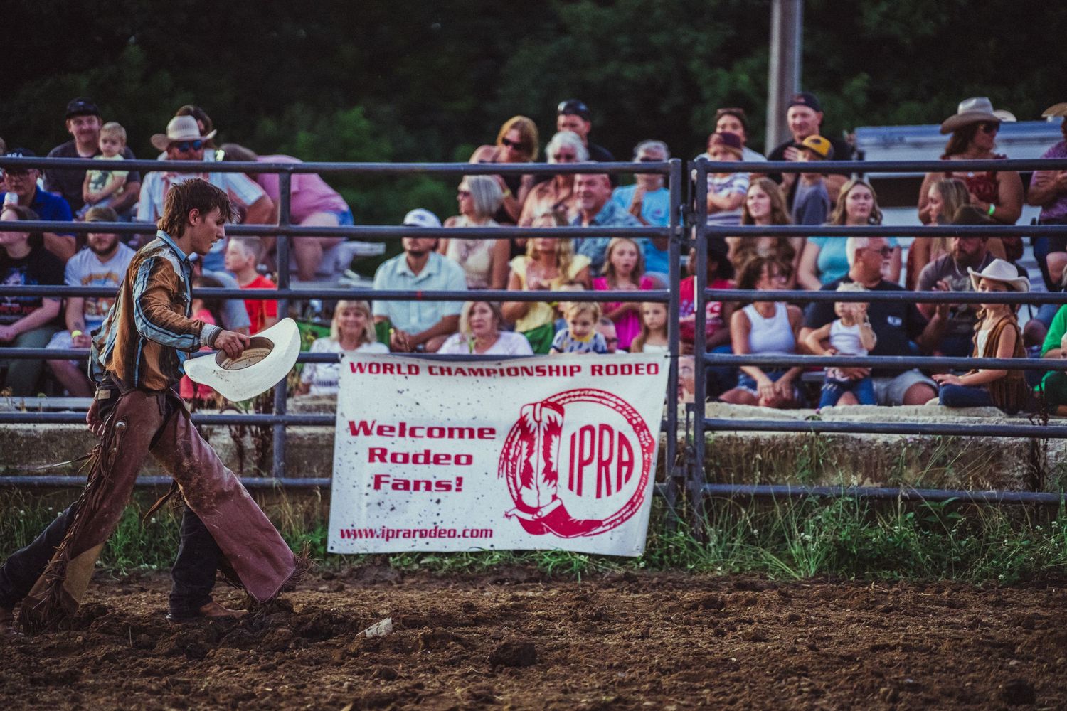Cowboy walks past IPRA World Championship Rodeo banner welcoming fans, with crowded grandstands in background.