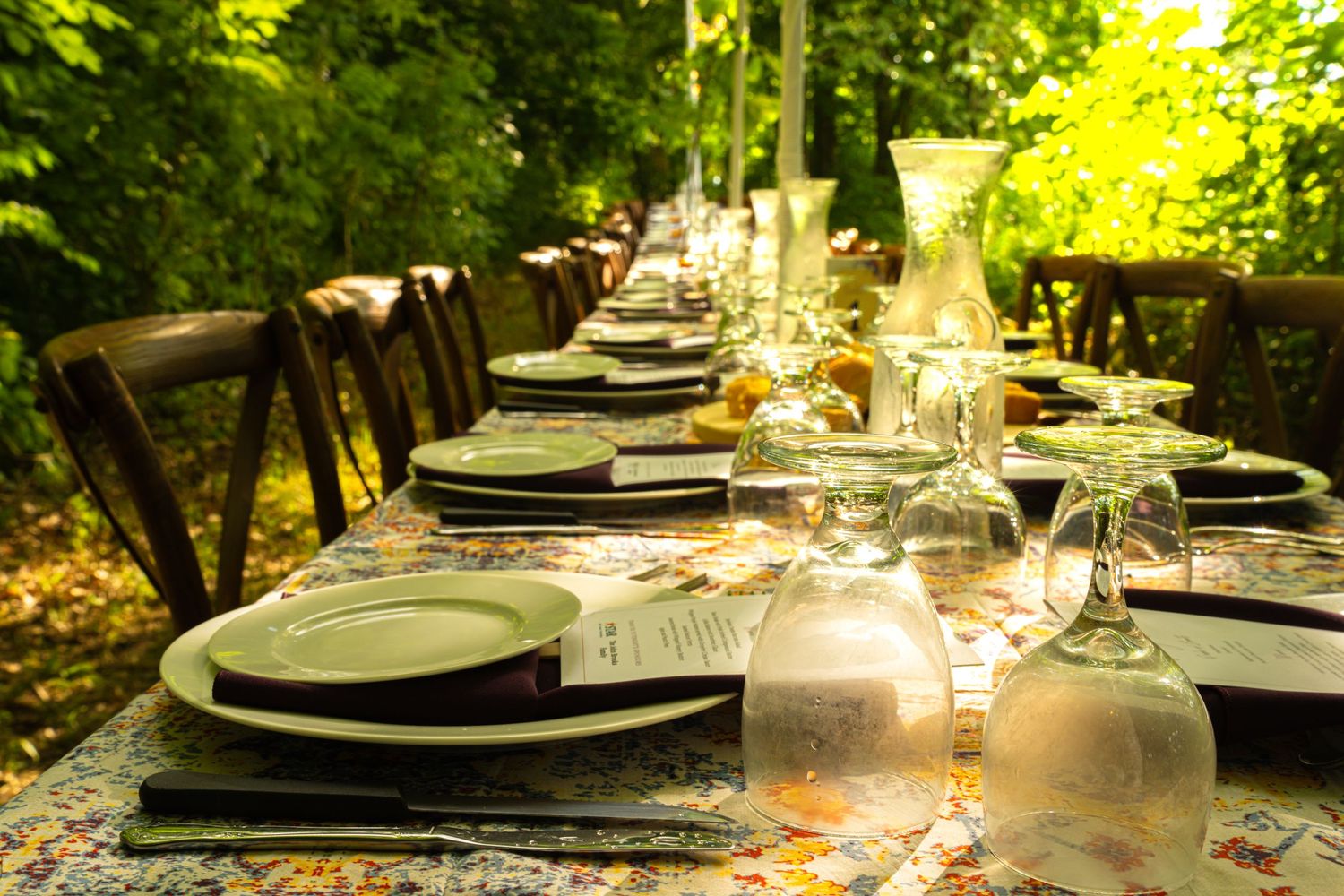 Elegant outdoor dining table set with plates, glassware, and menus surrounded by lush green trees.