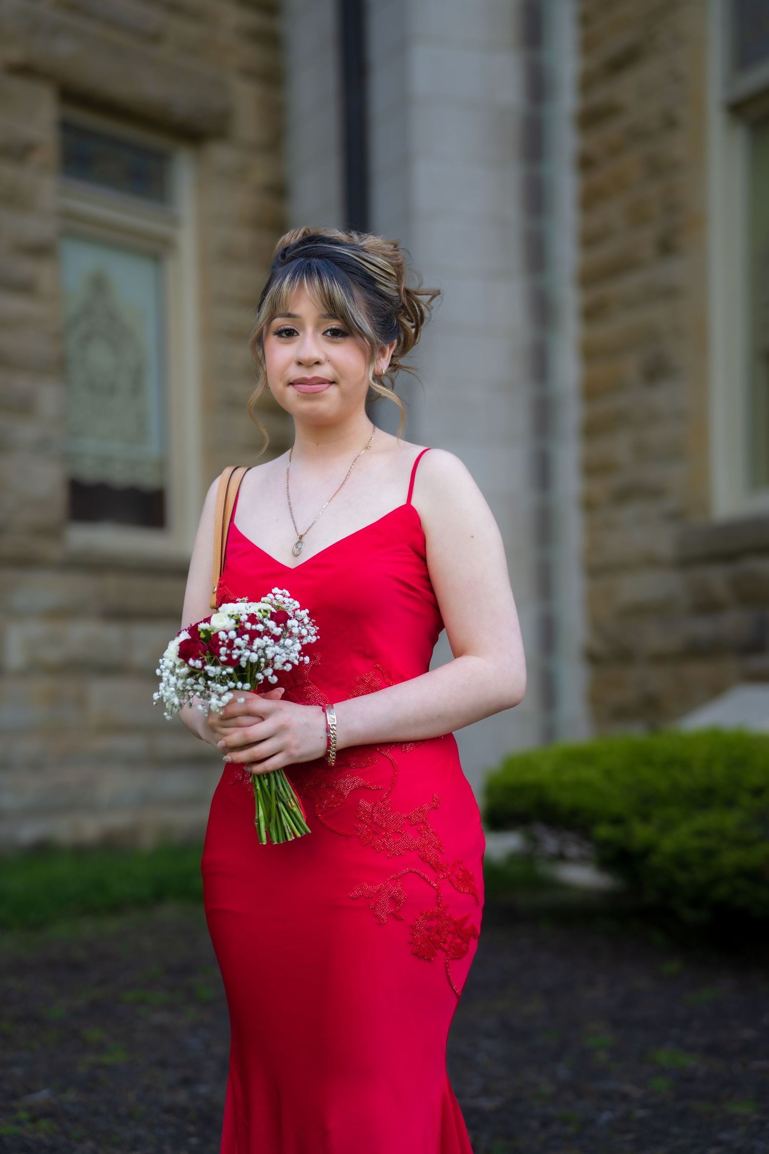 Young woman in red spaghetti strap gown holds red and white bouquet outside stone building.