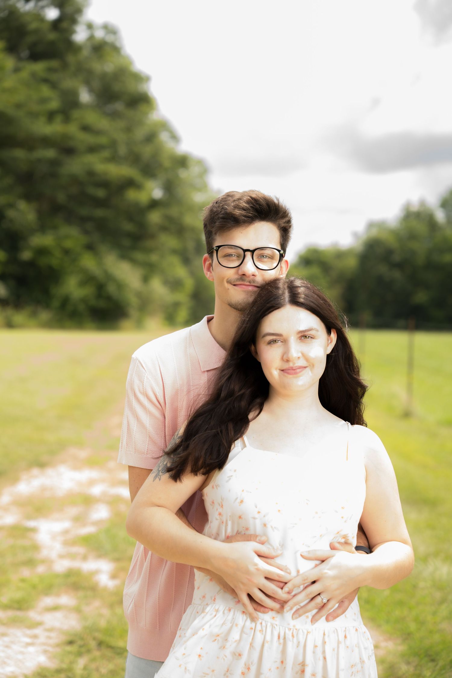 A young couple embracing outdoors, man with glasses hugging pregnant woman in white dress on a green field path.