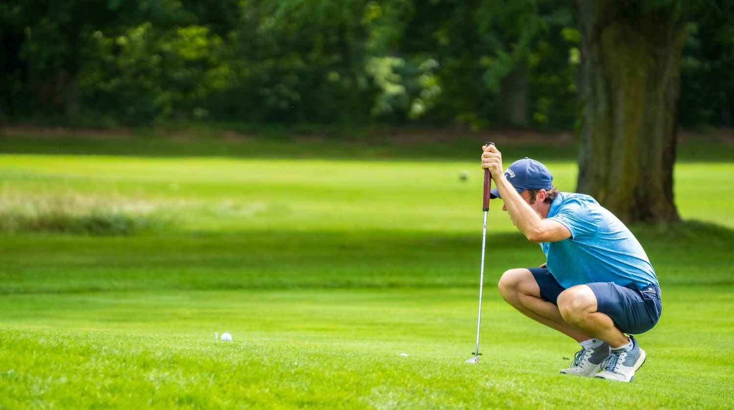 Golfer crouching on lush green fairway, lining up a putt with flag stick on sunny golf course.