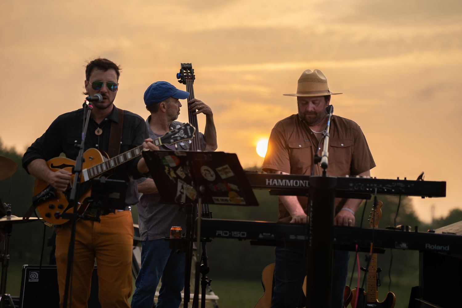 Three-piece band performing at outdoor sunset concert with guitar, bass, and Hammond organ.