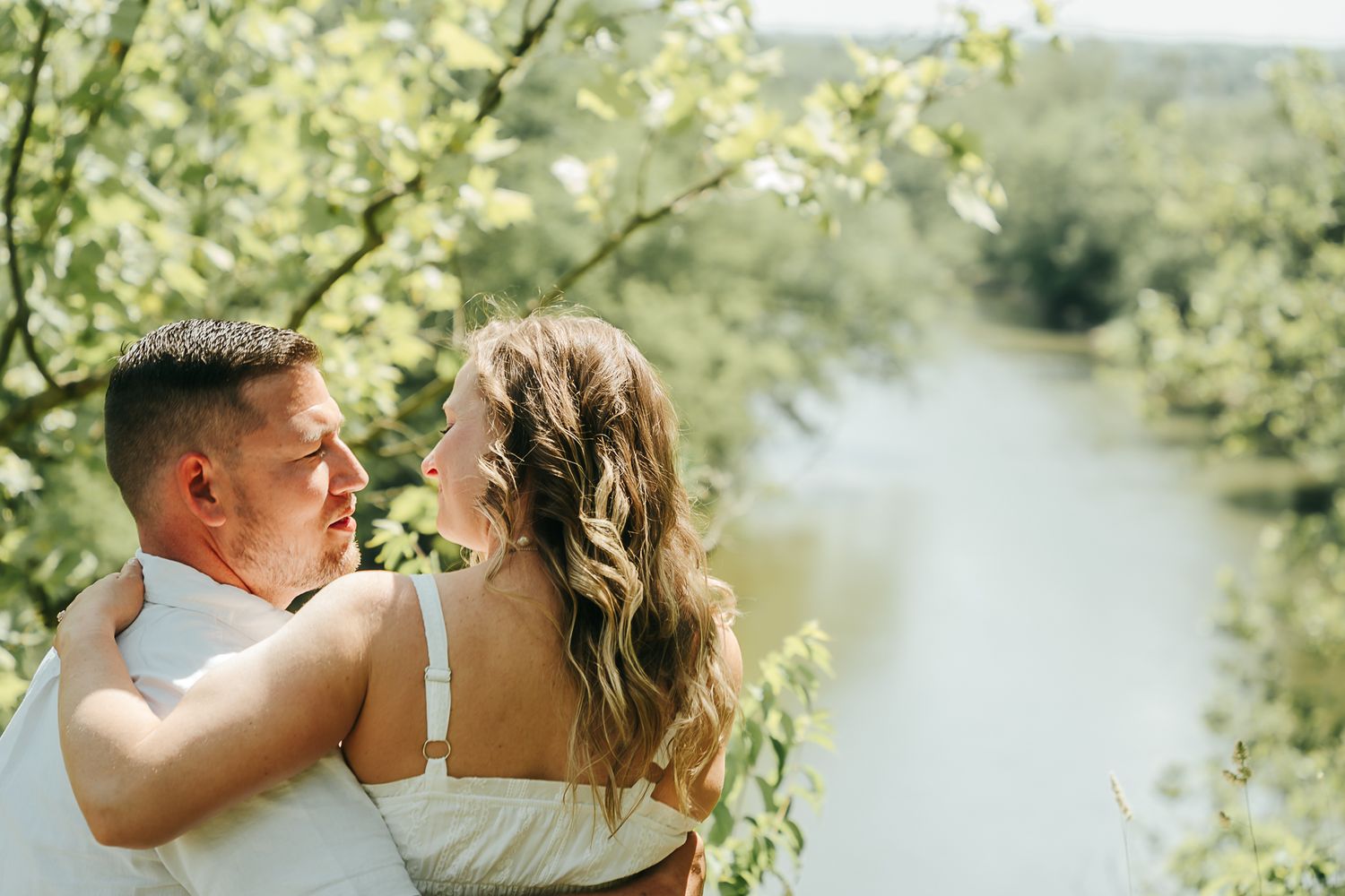 Couple embracing and gazing at each other with a serene river and green trees behind them.