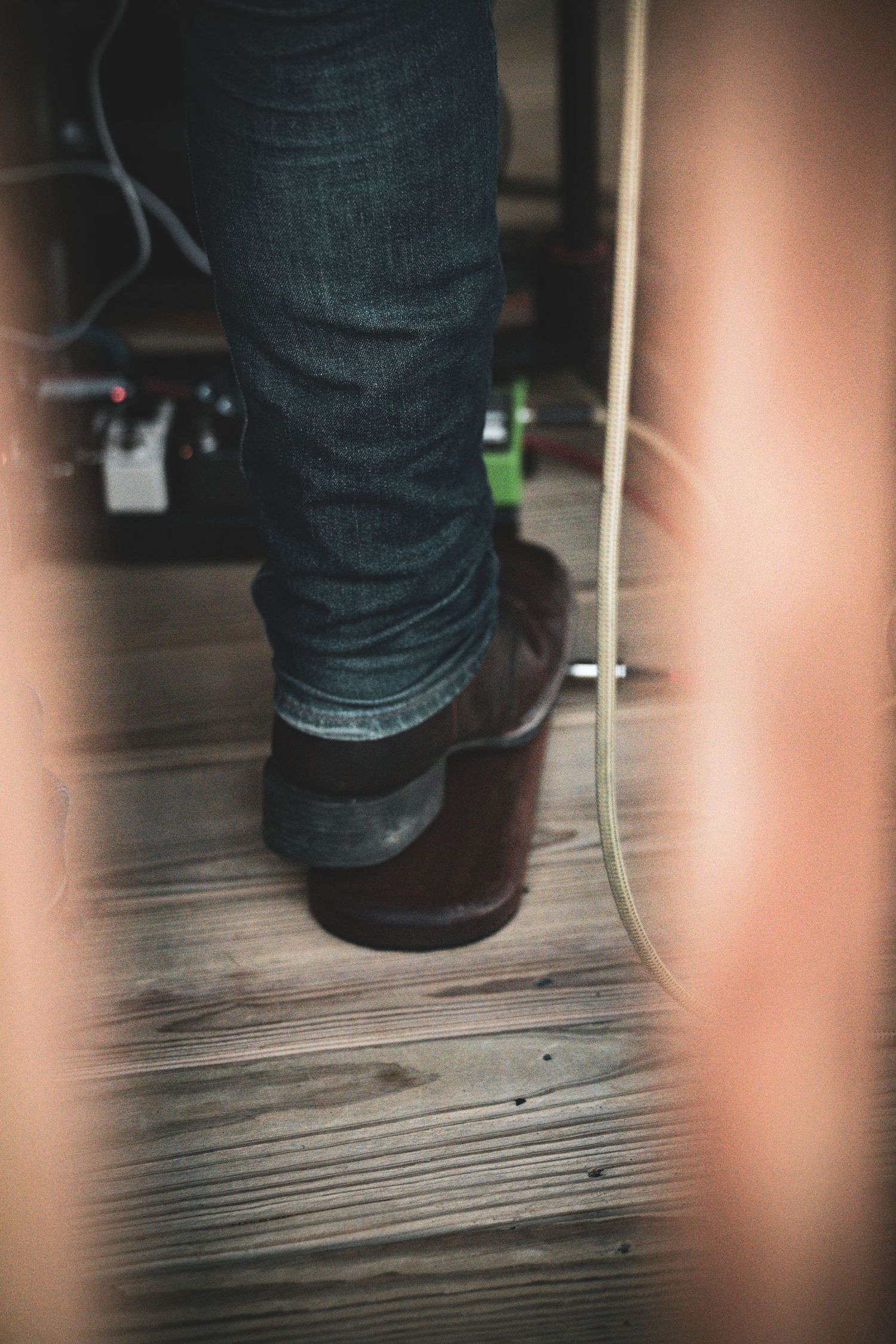 Close-up of musician's boots and jeans standing on wooden stage floor with cables and pedals visible.