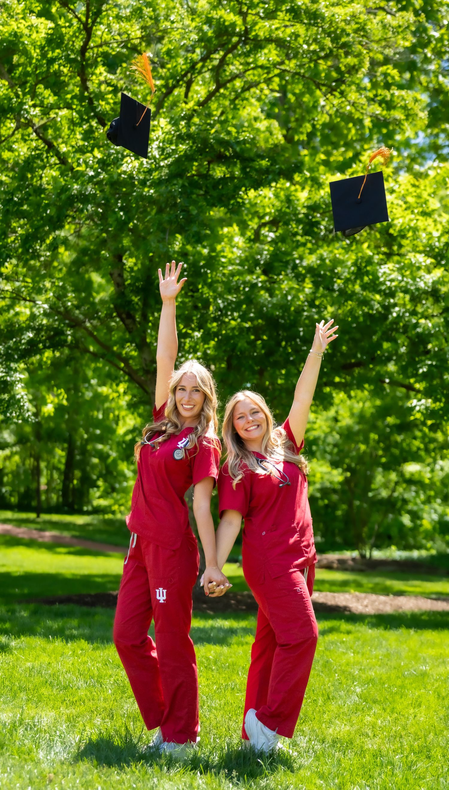 Two nursing graduates in red scrubs tossing mortarboard caps in the air on green campus lawn.