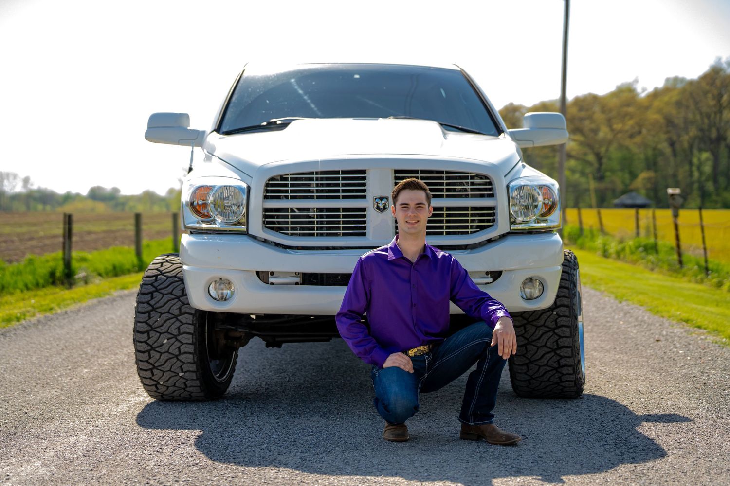 Teen boy in purple shirt crouching in front of lifted white Dodge Ram truck on a gravel road.