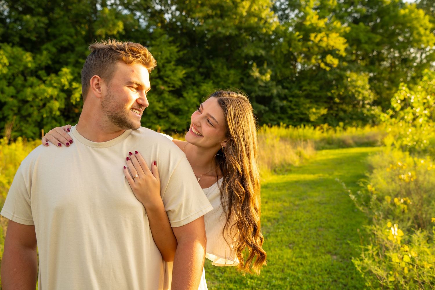 Happy engaged couple smiling at each other on a sunny path surrounded by green trees.
