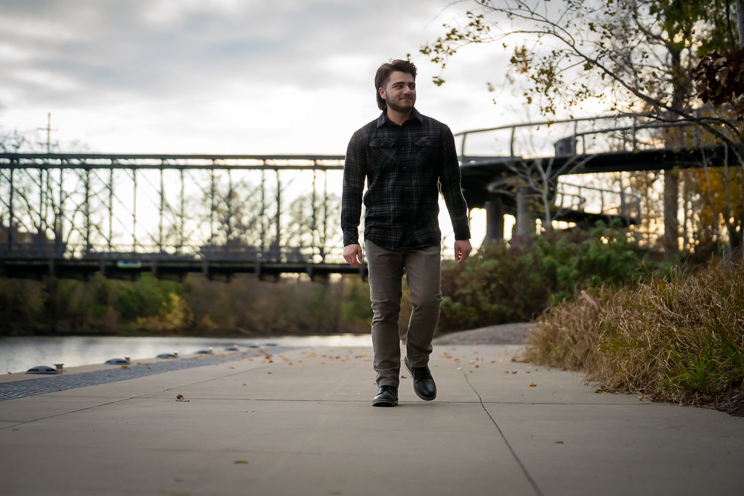 Smiling man in plaid shirt walking along riverside path with bridge in autumn background.