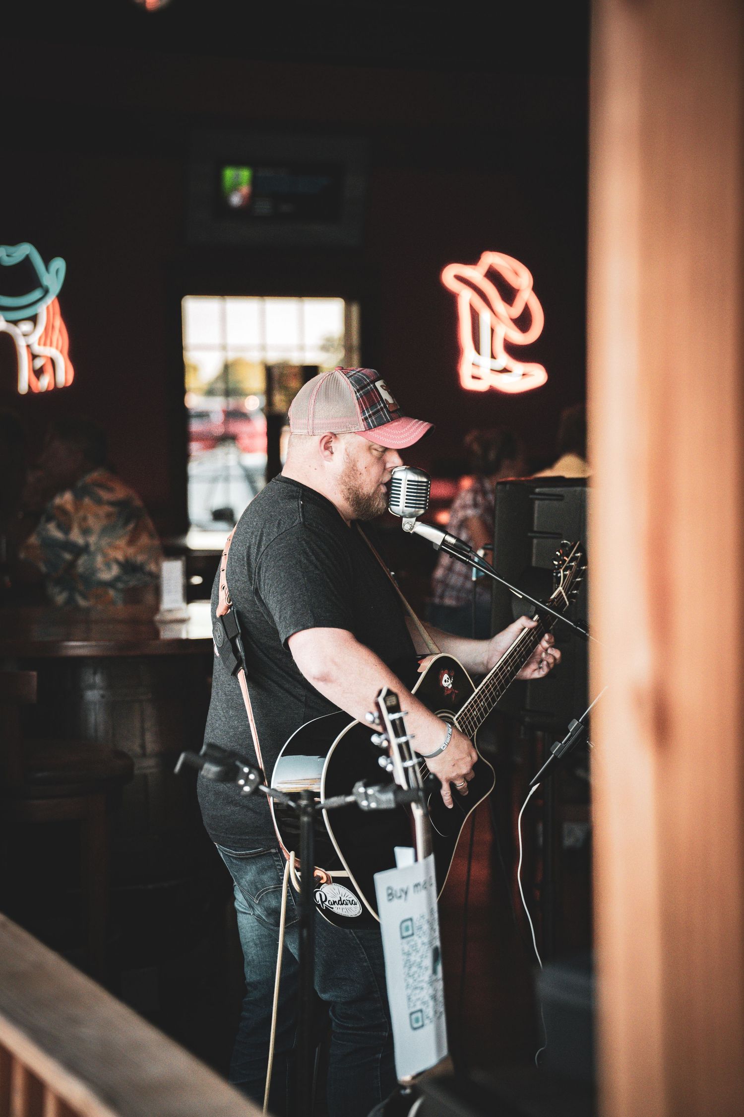 Country musician in black shirt and plaid cap plays black acoustic guitar inside bar with neon signs glowing.