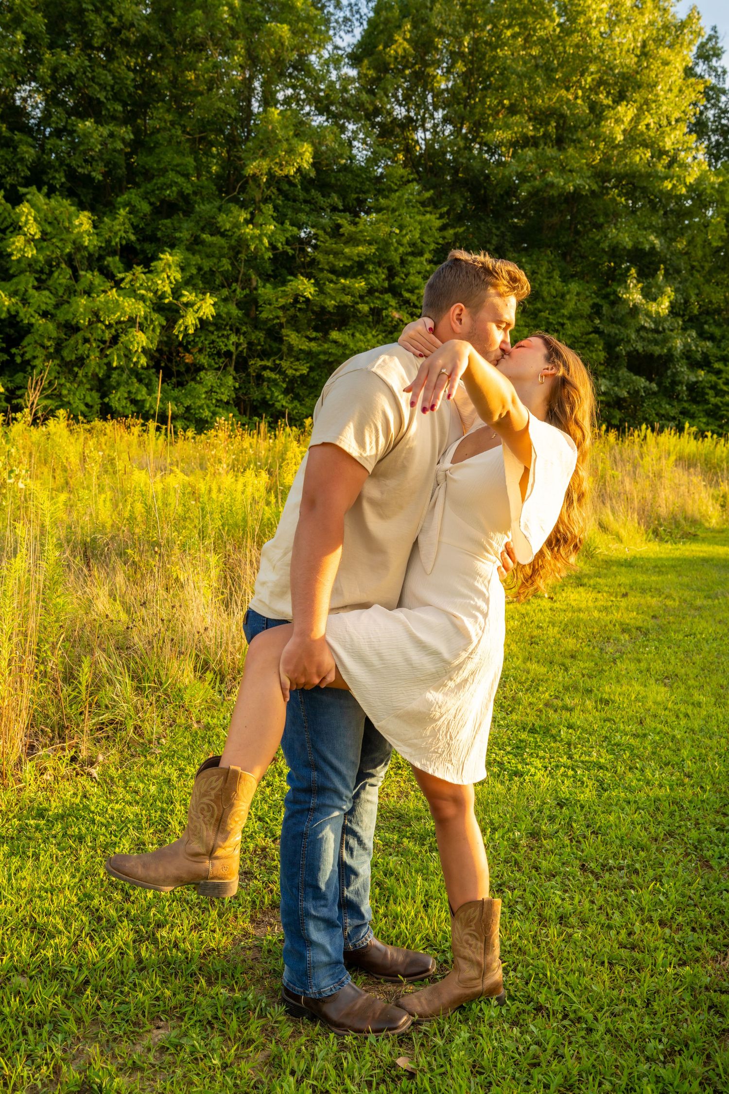 Couple sharing a romantic kiss in a golden sunlit meadow with lush green trees in the background.