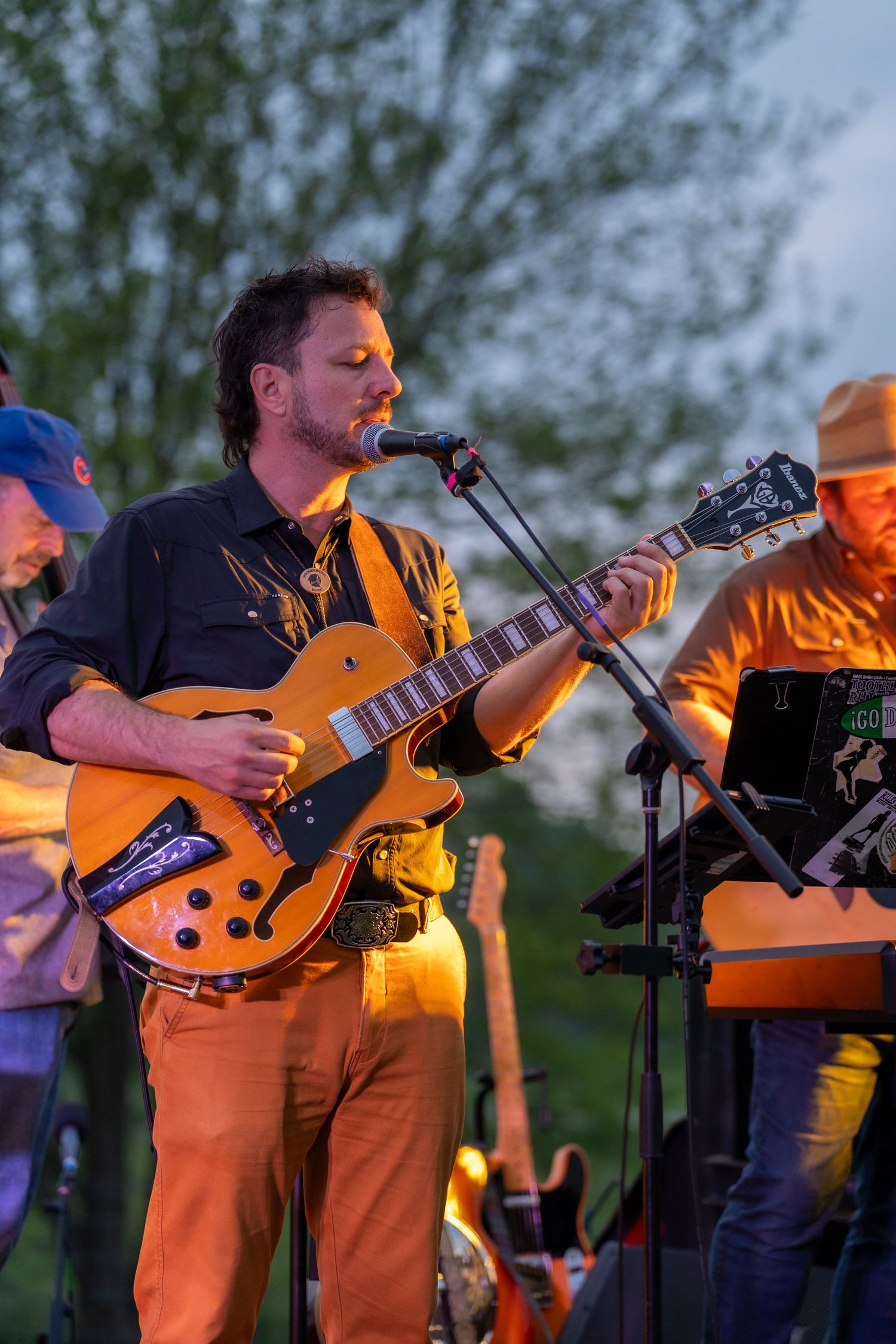 Male guitarist singing while playing archtop electric guitar at outdoor evening concert.