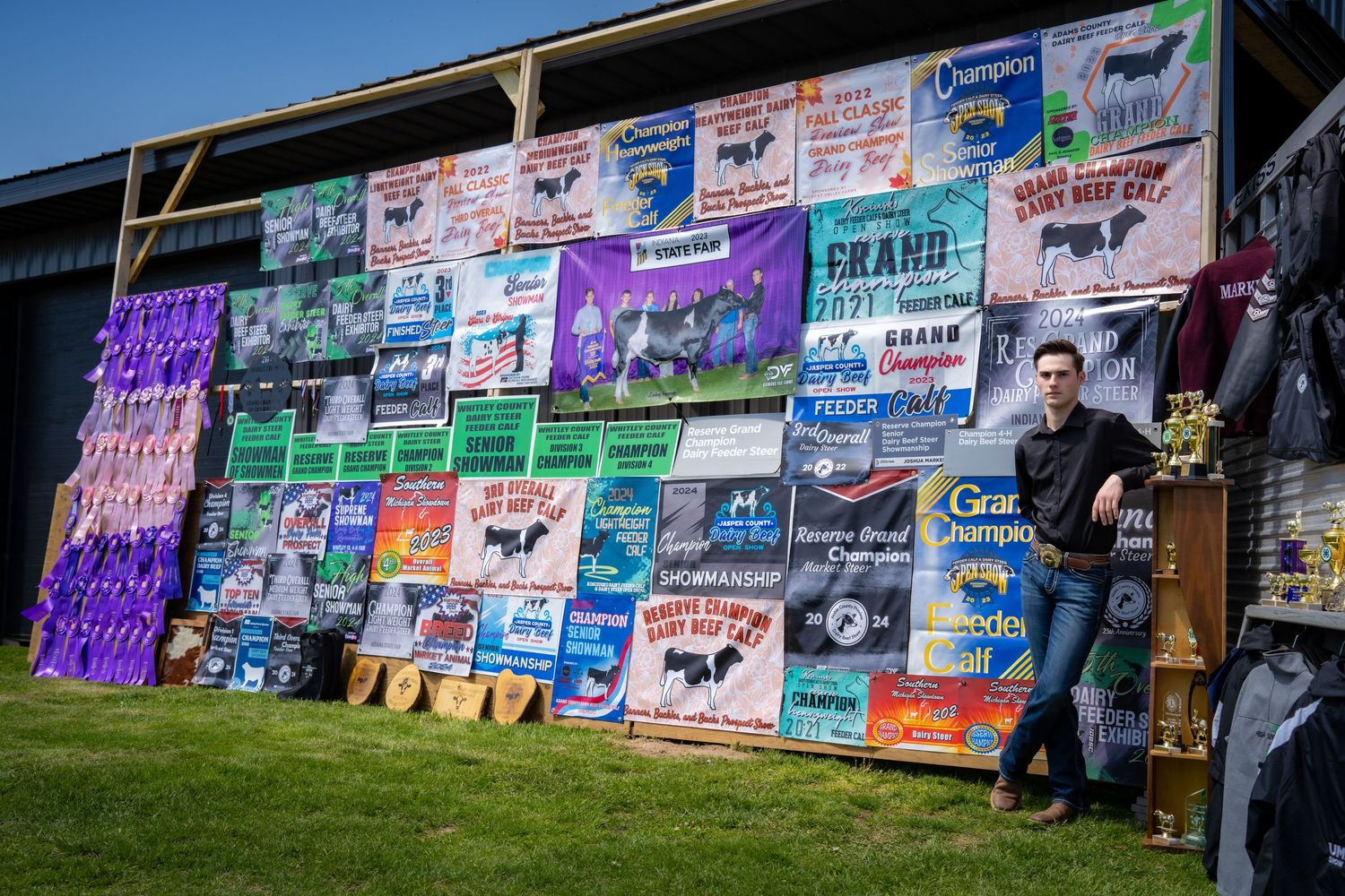 Young man stands beside colorful livestock show banners and championship signs displayed on a barn wall.