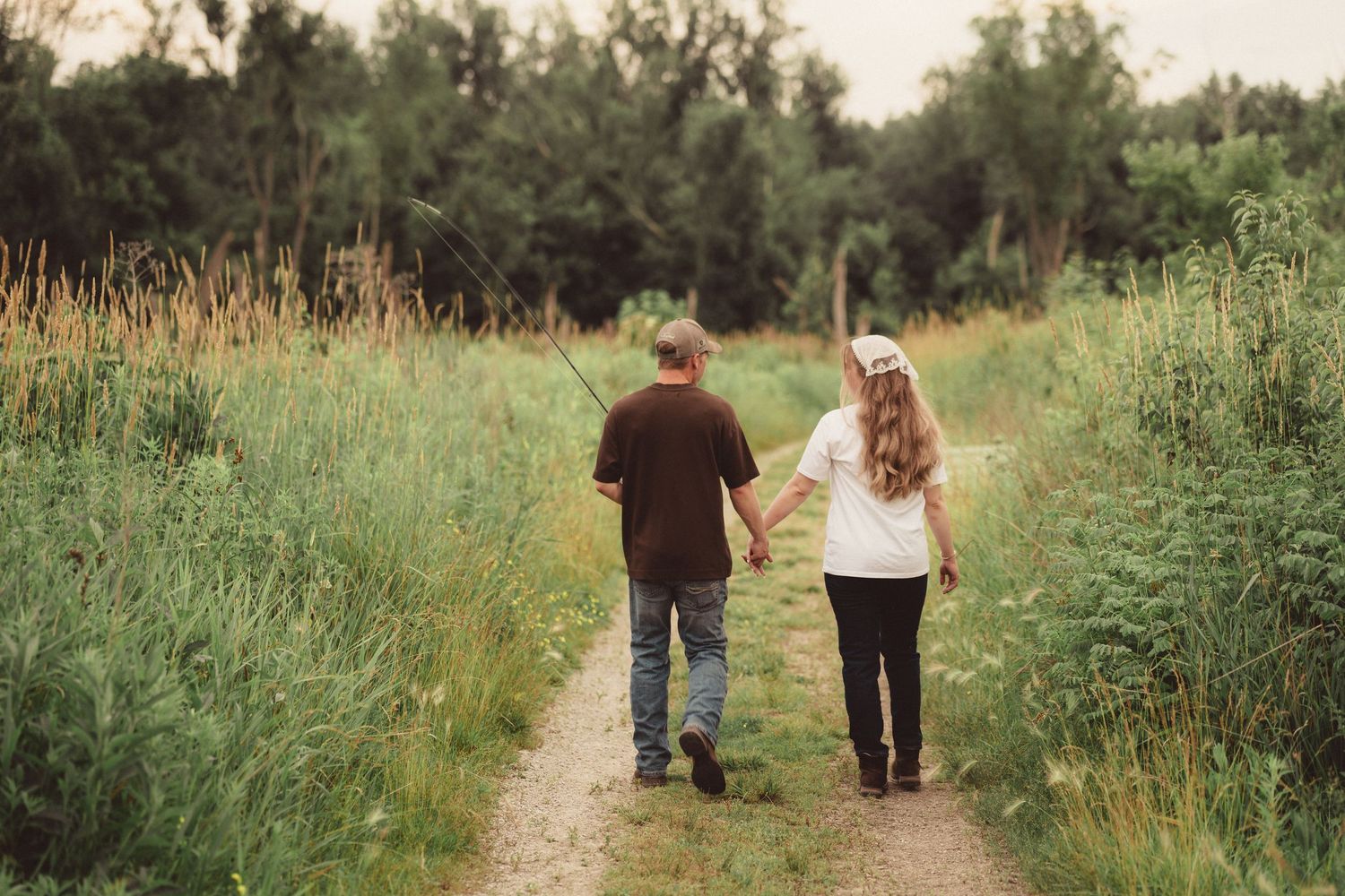 Couple walking hand in hand down a narrow path through tall green meadow grass.