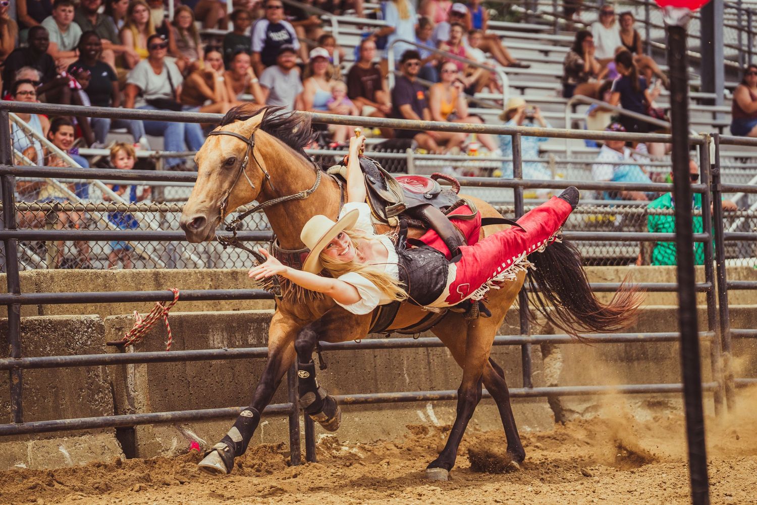 A rodeo rider struggles to stay on a bucking horse in a sandy arena with a crowd of spectators watching.
