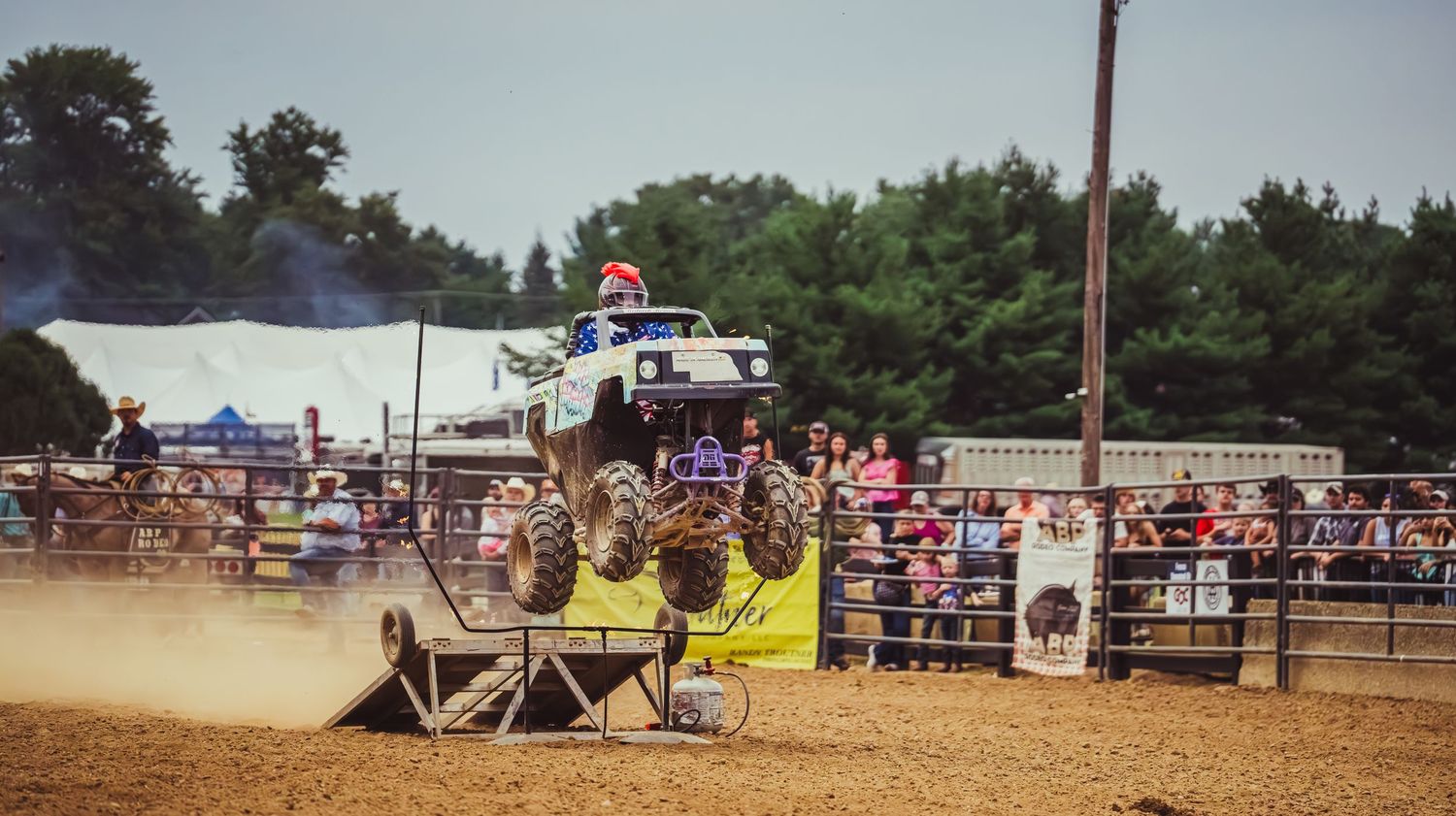 Monster truck soaring over a dirt ramp at an outdoor arena with a crowd of spectators watching the stunt show.