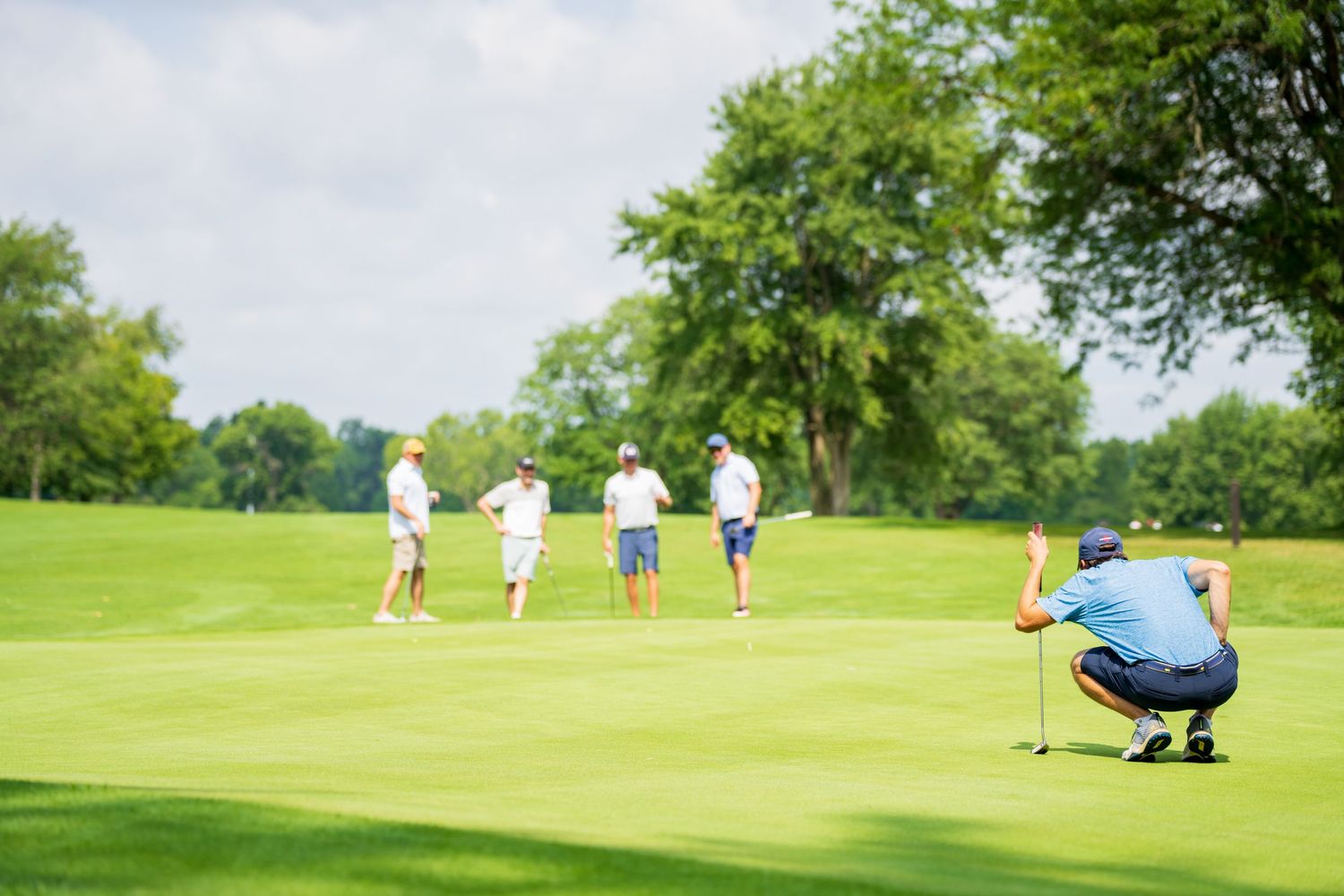 Golfer crouching to read the green while three players stand in background on sunny golf course.