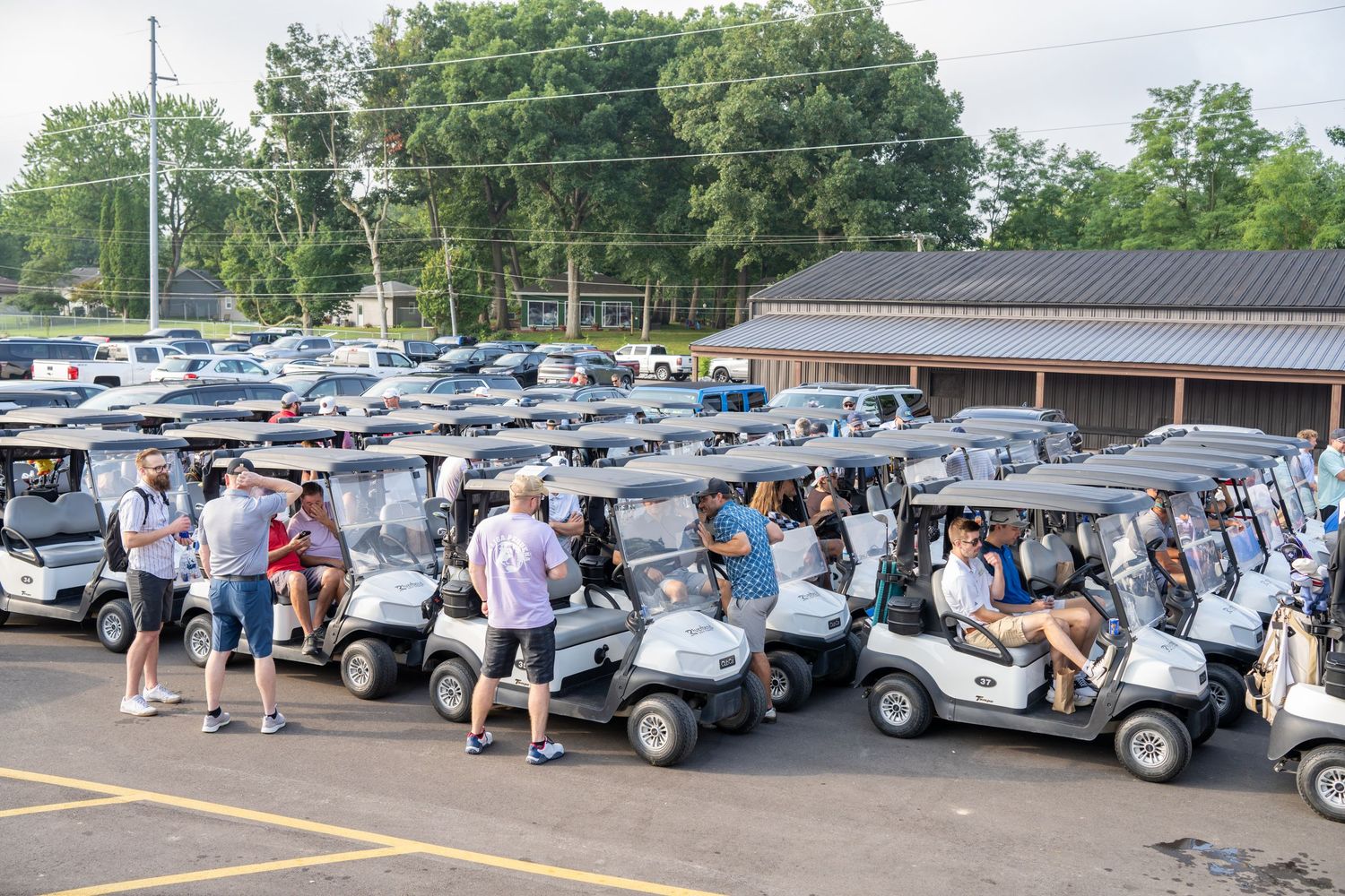 Large group of golfers gathering around white golf carts in a parking lot before a golf tournament.
