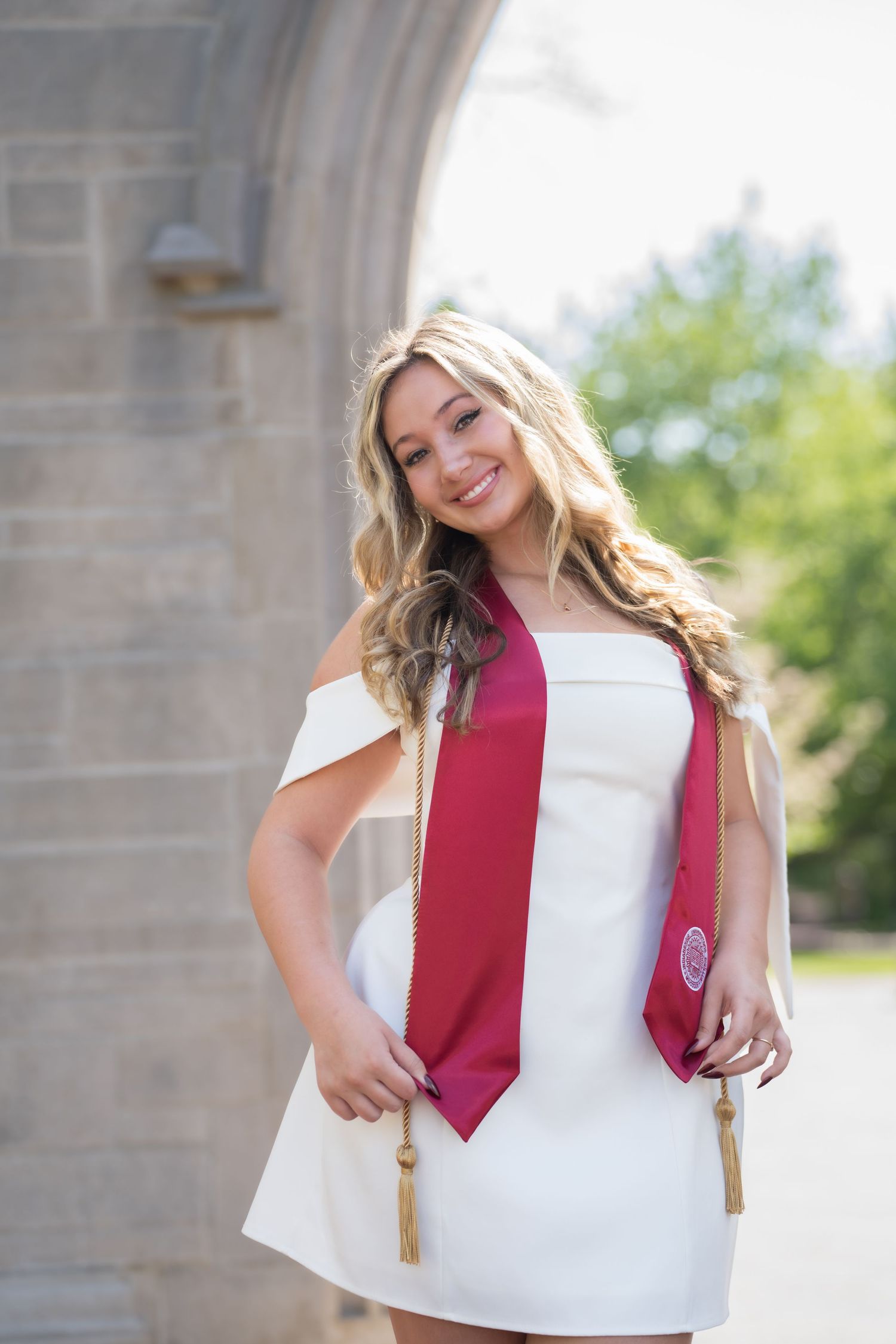Smiling blonde graduate in white dress with crimson graduation stole poses under stone arch outdoors.