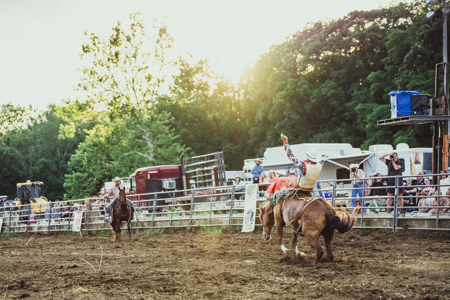 Cowboys compete in a thrilling rodeo event at a rustic outdoor arena surrounded by lush green trees in warm sunlight.