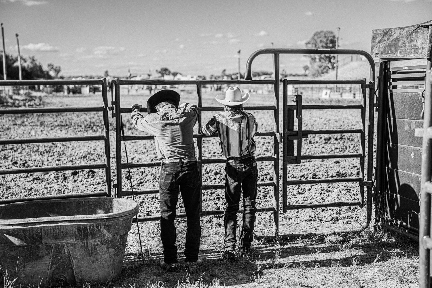 Two cowboys in hats work beside metal livestock gates on a ranch in this black and white rural western photo.