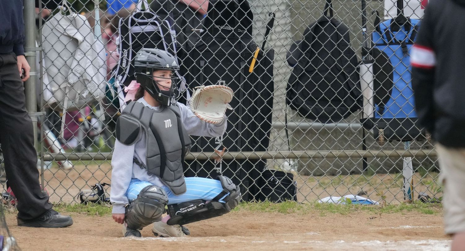 Baseball catcher in full gear crouching at home plate during an outdoor game near a chain-link fence.