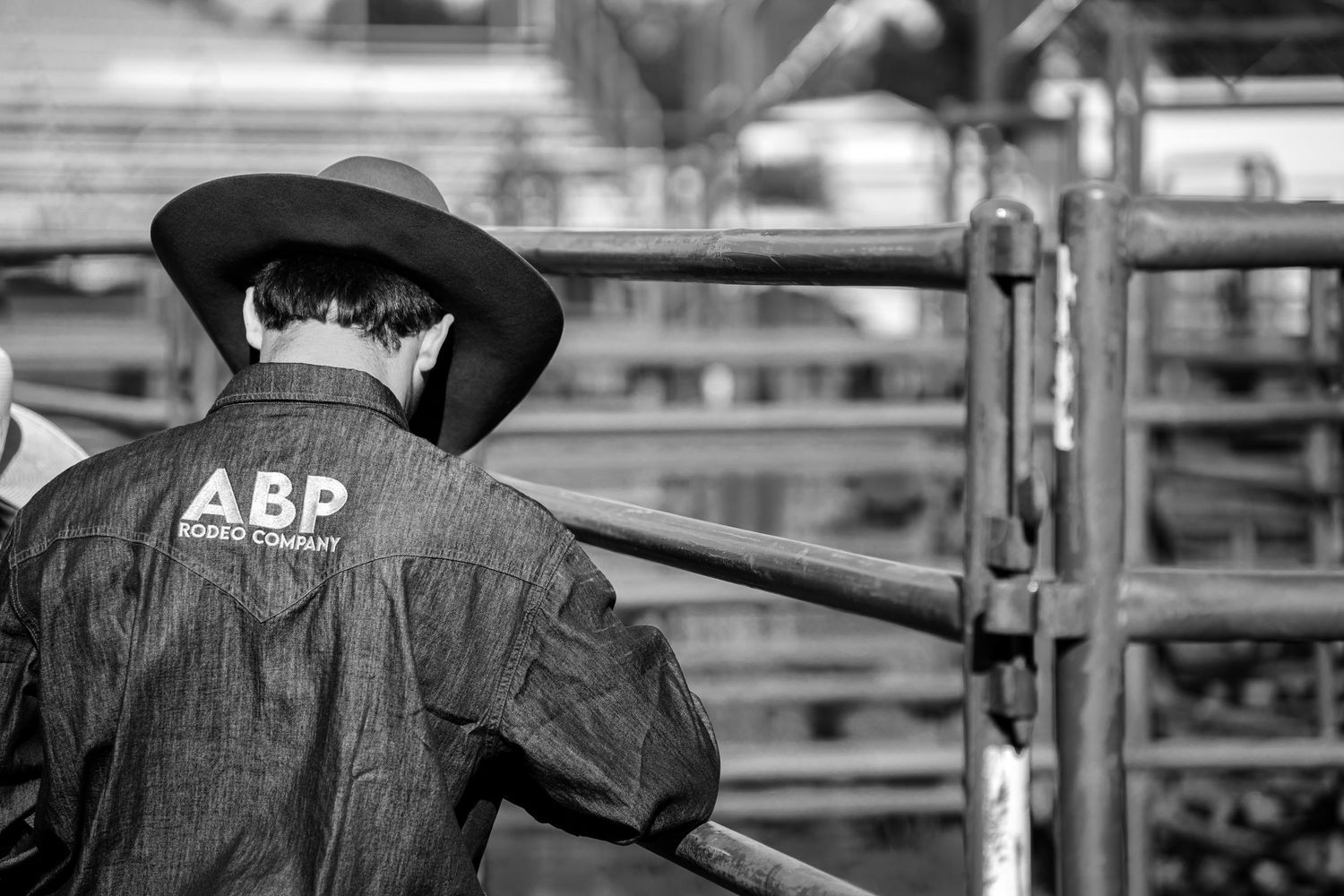Black and white photo of a cowboy in an ABP rodeo jacket leaning on a fence at a rodeo event.