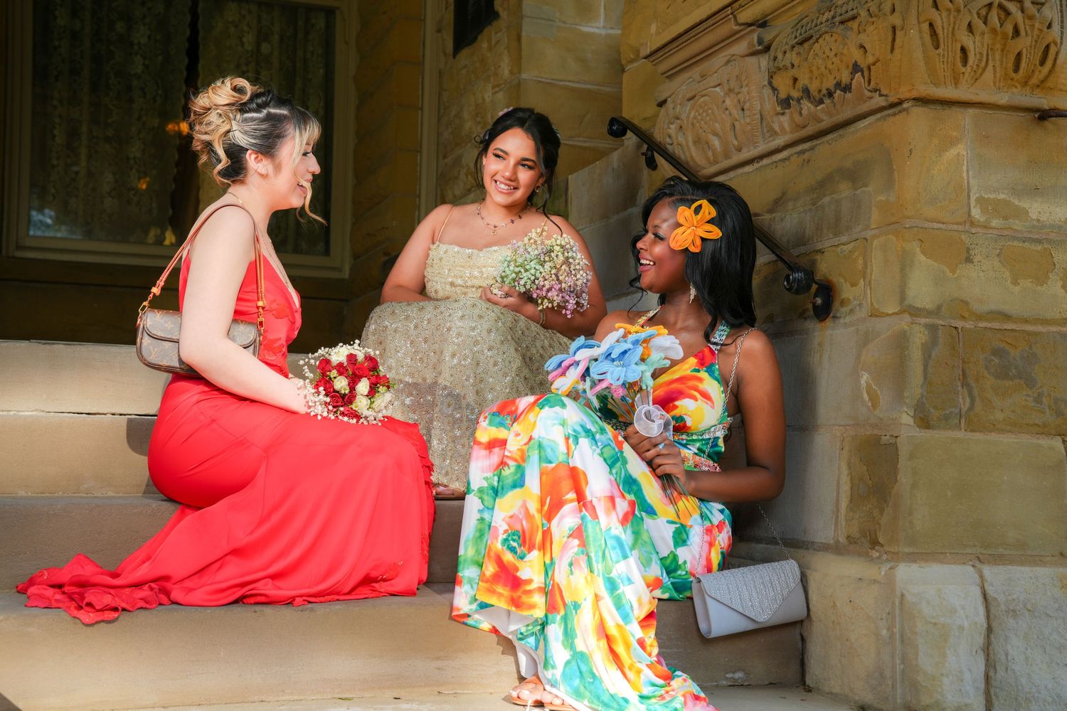 Three women in colorful formal dresses sit on stone steps, chatting and holding bouquets at a wedding.