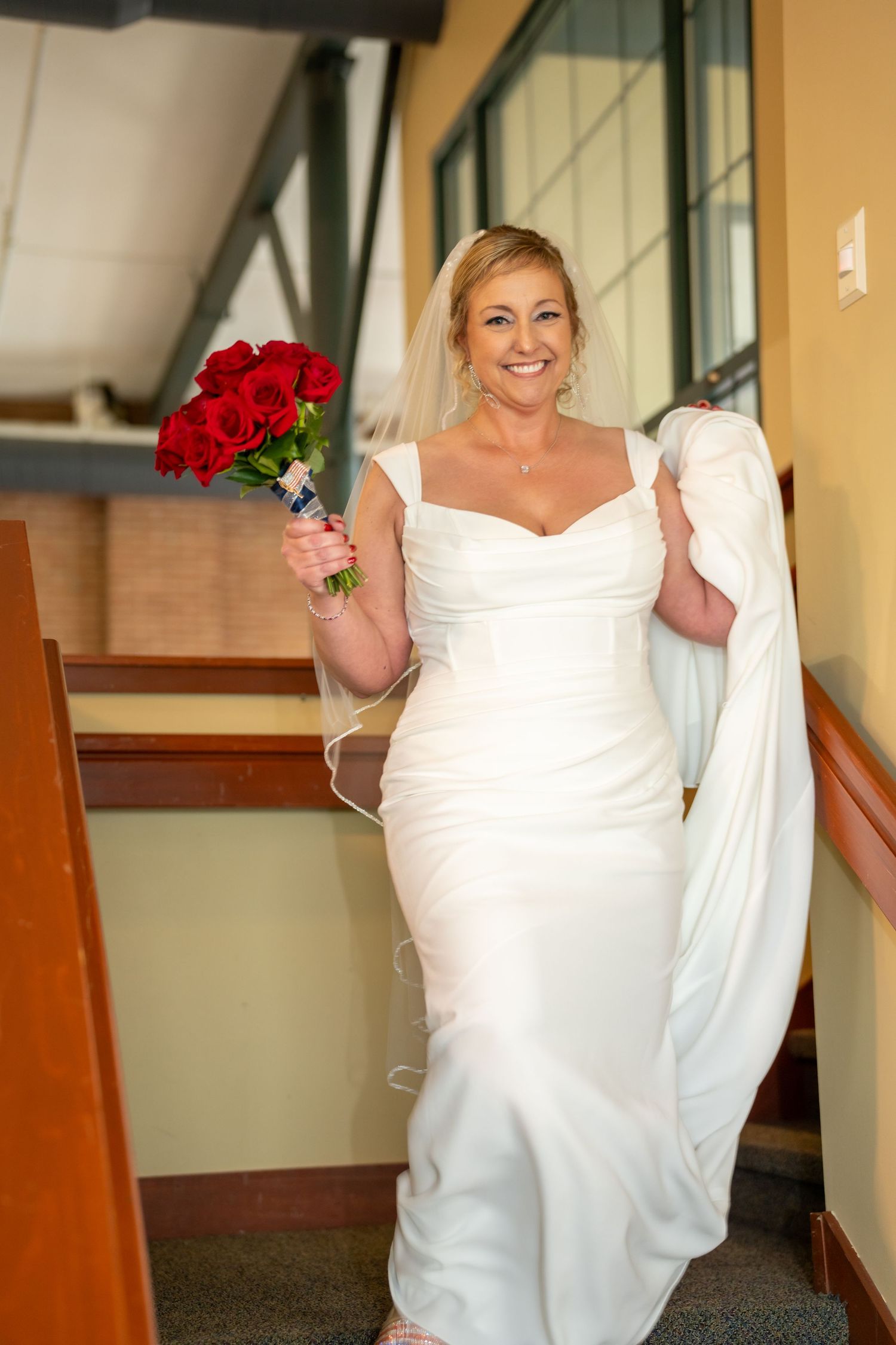 Smiling bride in white gown holding red roses bouquet, walking up stairs with veil trailing behind her.