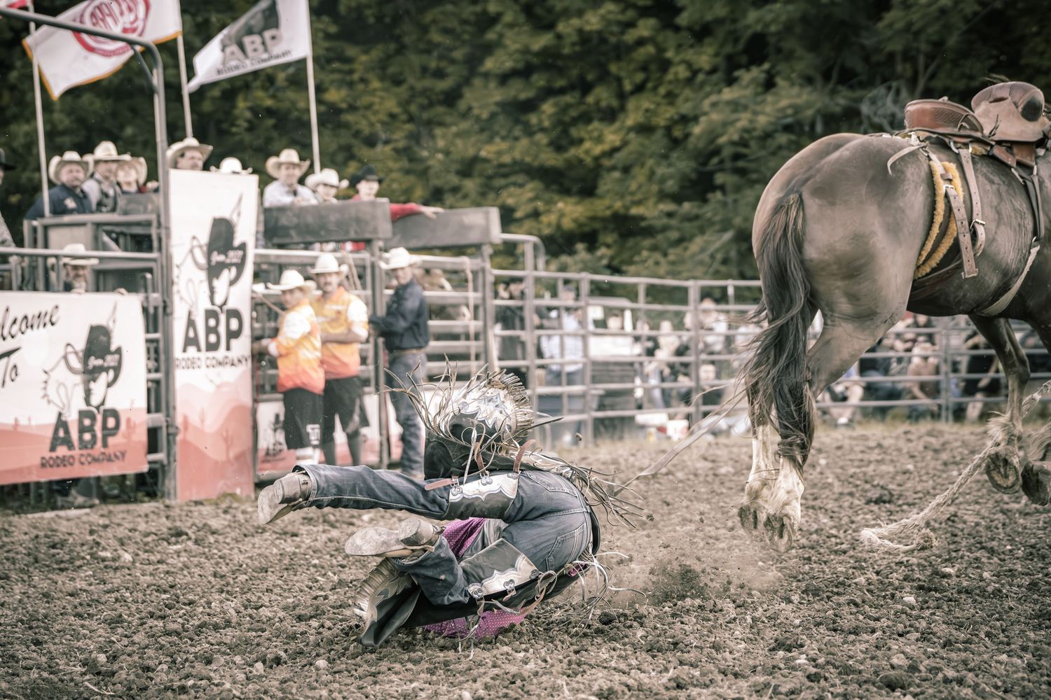 Fallen rodeo rider on the dirt ground next to a horse at an ABP rodeo event with crowd watching.